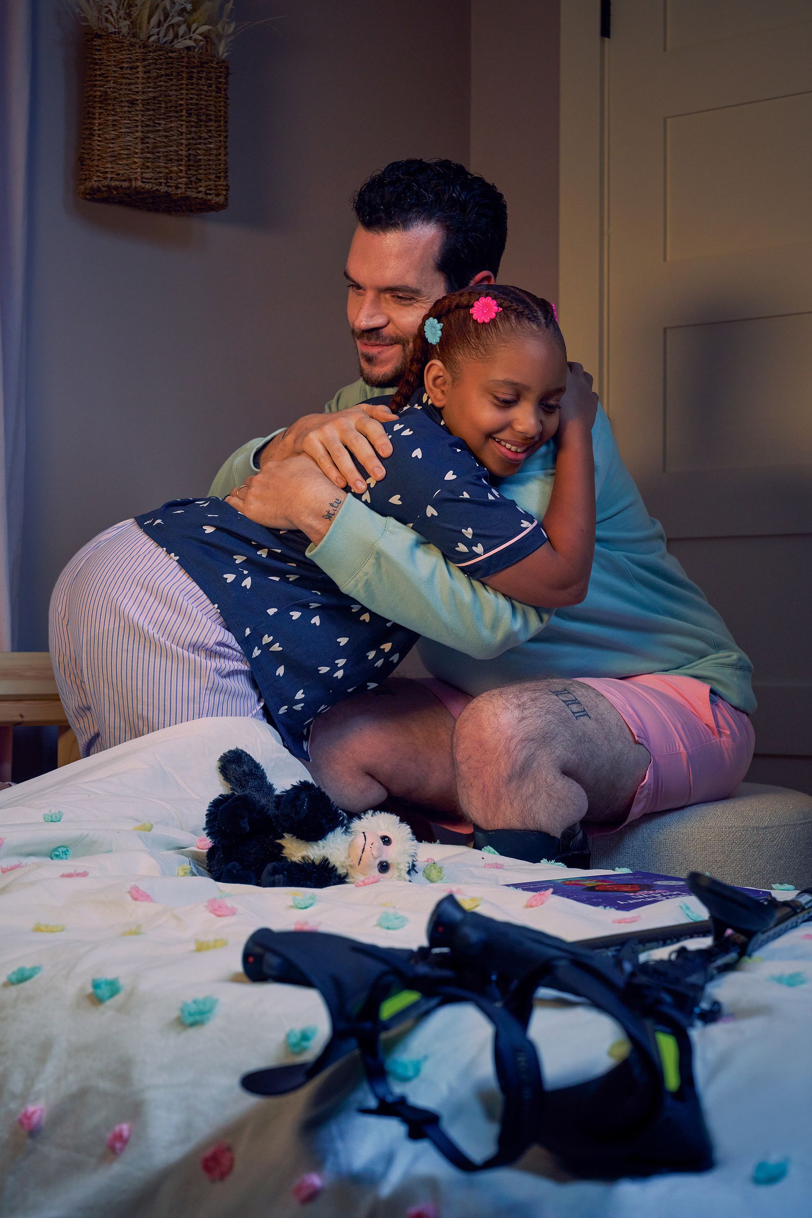 A father and daughter smile and embrace in her bedroom at night. The father is a white man with Cerebral Palsy who wears leg braces. His black forearm crutches lie on the foot of the daughter's bed. The daughter is a Black multiracial American girl with thick braided red hair adorned with two flower barrettes and is wearing blue patterned pajamas. A Capuchin monkey plushie lays on her bed.