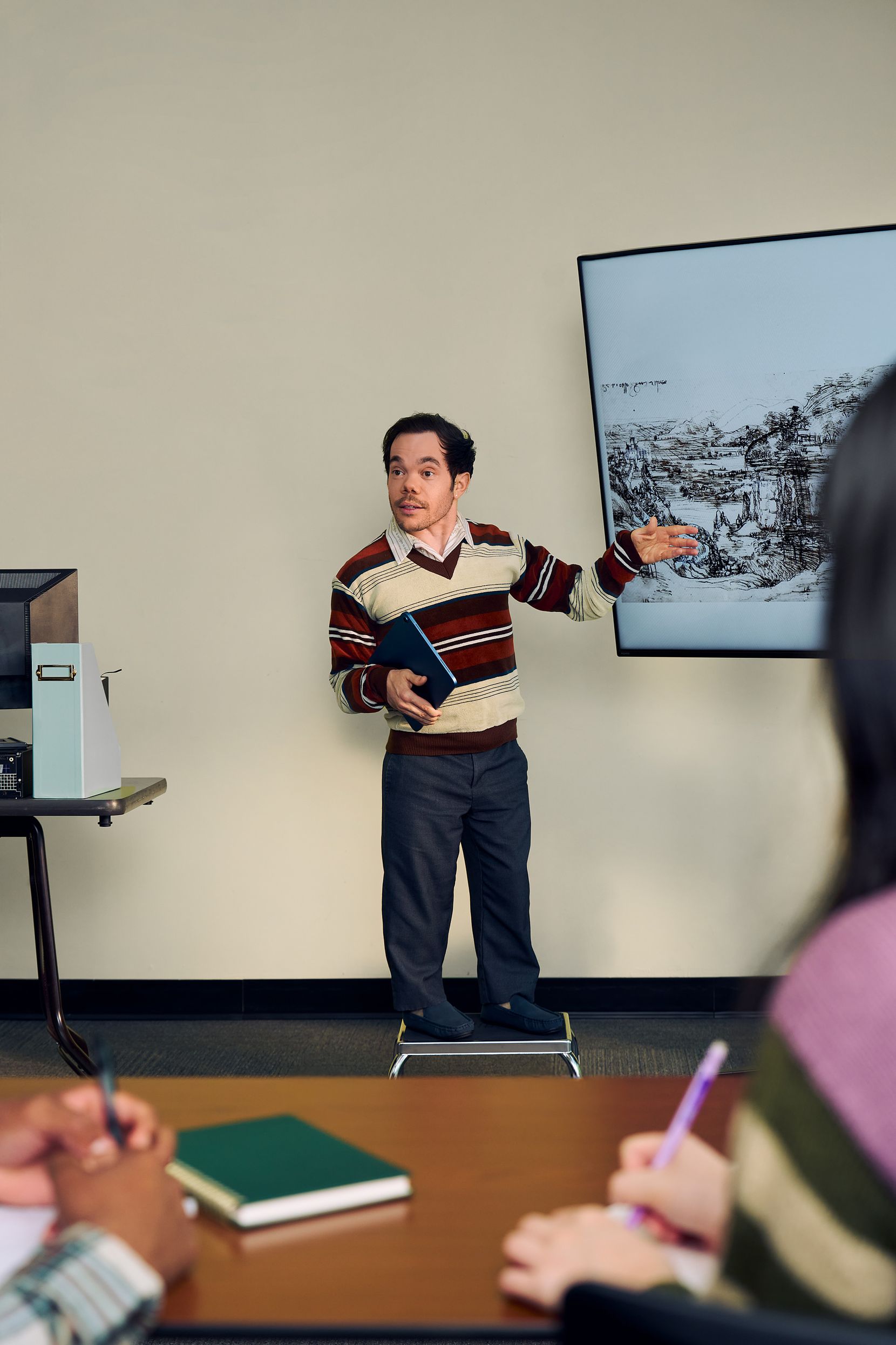 A white male professor with medium brown hair and light brown eyes who has dwarfism stands on a stool and gestures at a large monitor next to him, displaying a pen and ink drawing by Leonardo da Vinci. He is holding a tablet in his other hand. He has short brown and is wearing a red striped sweater, a collared shirt, and dark dress pants. In front of him, two students write in notebooks.