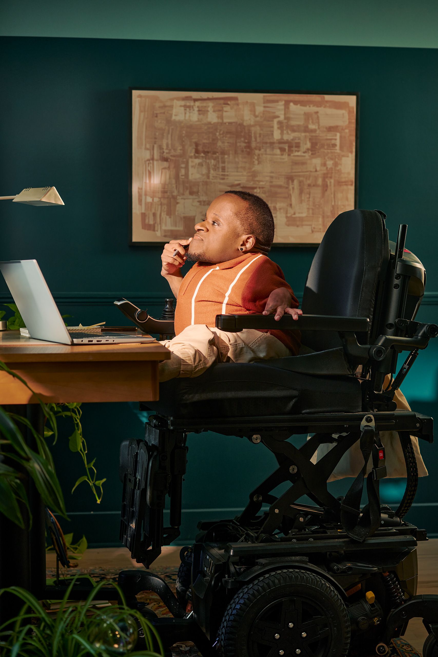 A black man who is a little person is seated in his power wheelchair in a dark teal home office. He holds a pen in his hand up to his chin and looks away, deep in thought. A laptop, notebook, pen, and lamp are on the desk in front of him. His wheelchair is in an elevated position, allowing him to be level with his desk. His desk is surrounded by several house plants and an abstract art piece. He has closely cropped black hair, brown eyes, and is wearing a rust-orange knit quarter-zip sweater with white vertical stripes, light khaki trousers, and a small stud earring. A tote bag hangs on the back of his wheelchair.