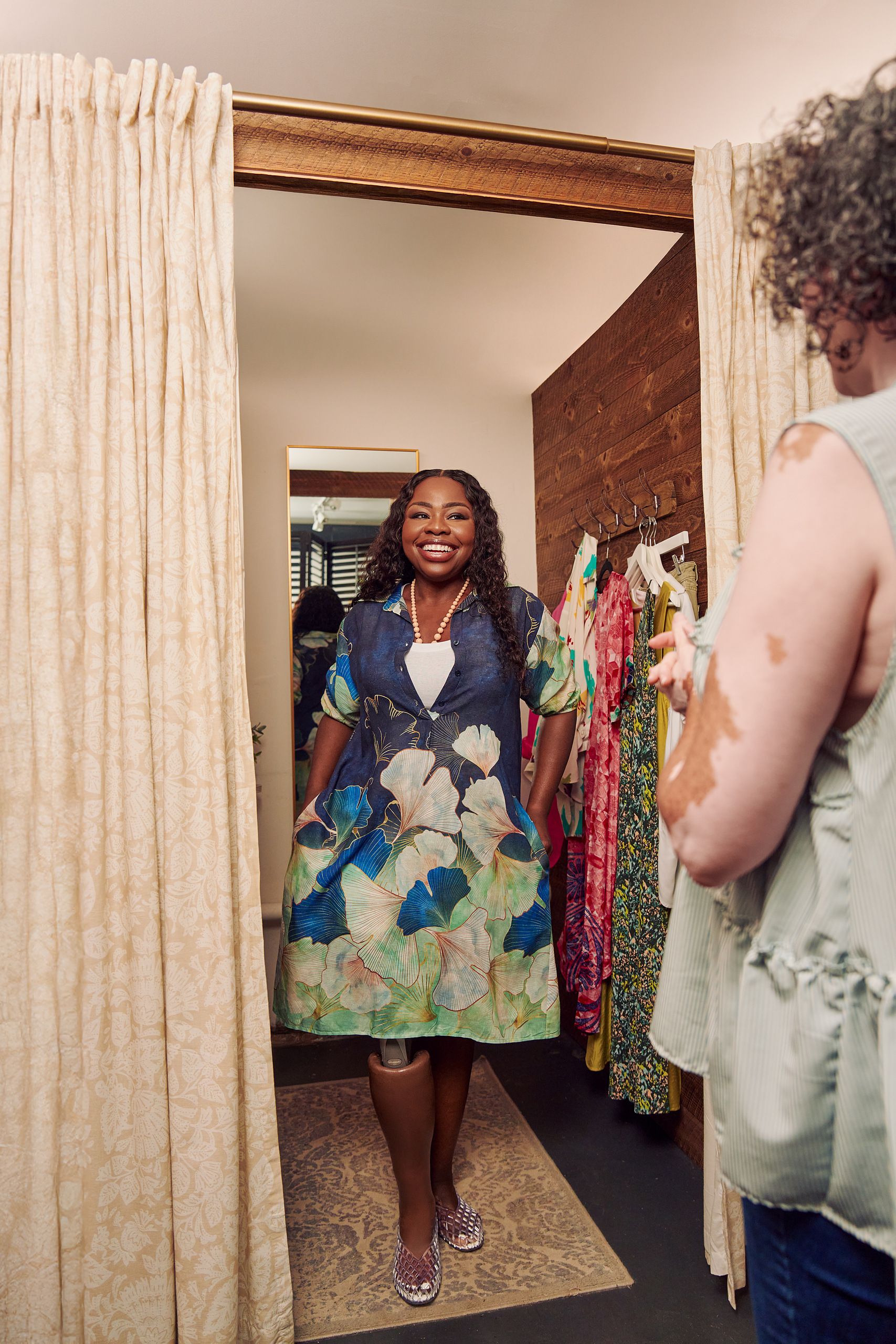 A Black woman with a prosthetic leg poses in a dressing room of a boutique with a big smile on her face. She is wearing a blue patterned collared dress and jelly sandals. She has her hands in the pockets of the dress and looks at the shopkeeper, partially off-camera. Several high end garments are on hooks in the dressing room. The shopkeeper is a Black woman with Vitiligo. She has a short, curly bob and is wearing glasses, a flowy blouse, and jeans.