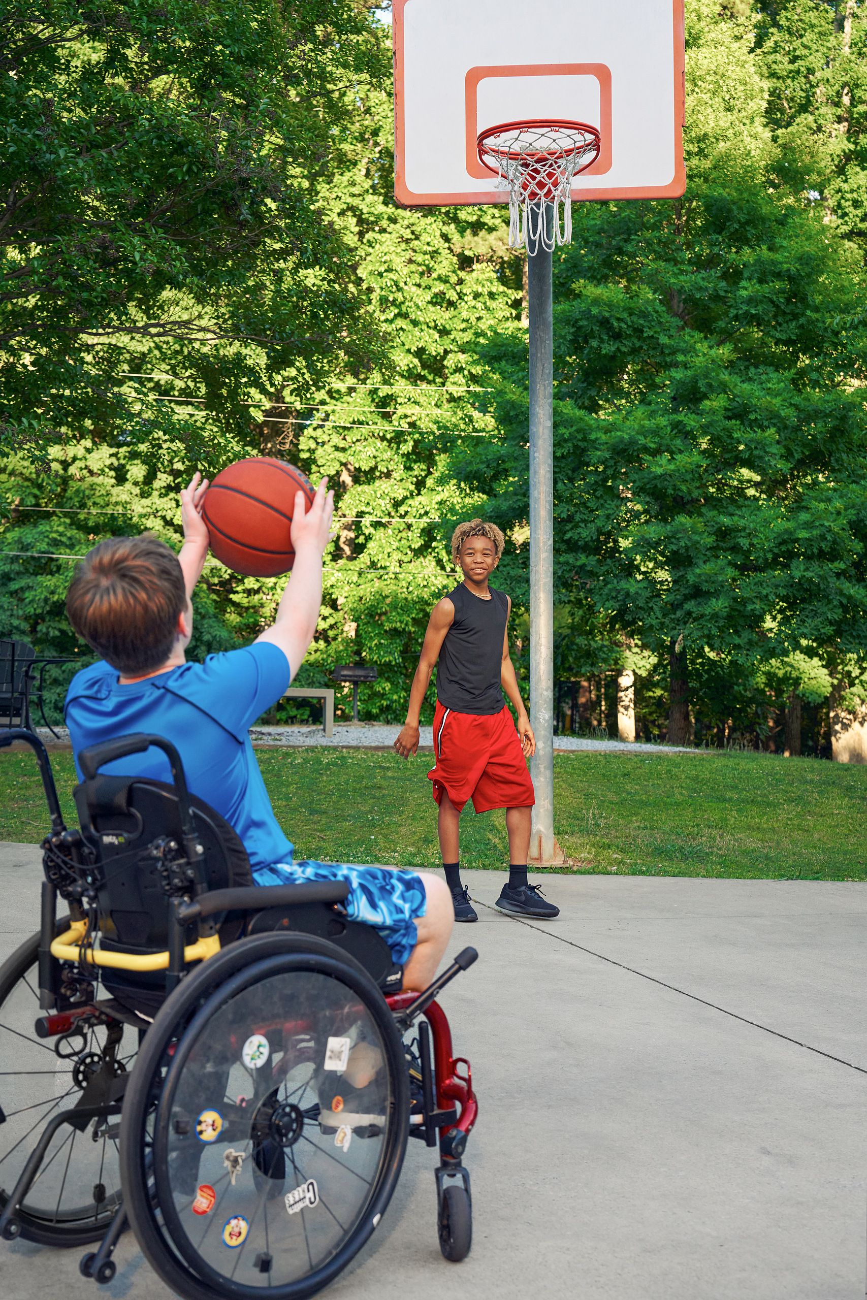 Two teens play basketball at a neighborhood park. One of the friends, a white boy seated in a manual wheelchair, passes the ball while a light-skinned African American male, who is standing near the hoop, watches. The teen seated in the manual wheelchair has short brown hair is wearing blue athletic wear and faces away from the camera. The African American teen has curly brown to blonde hair and is wearing black and blue athletic wear.