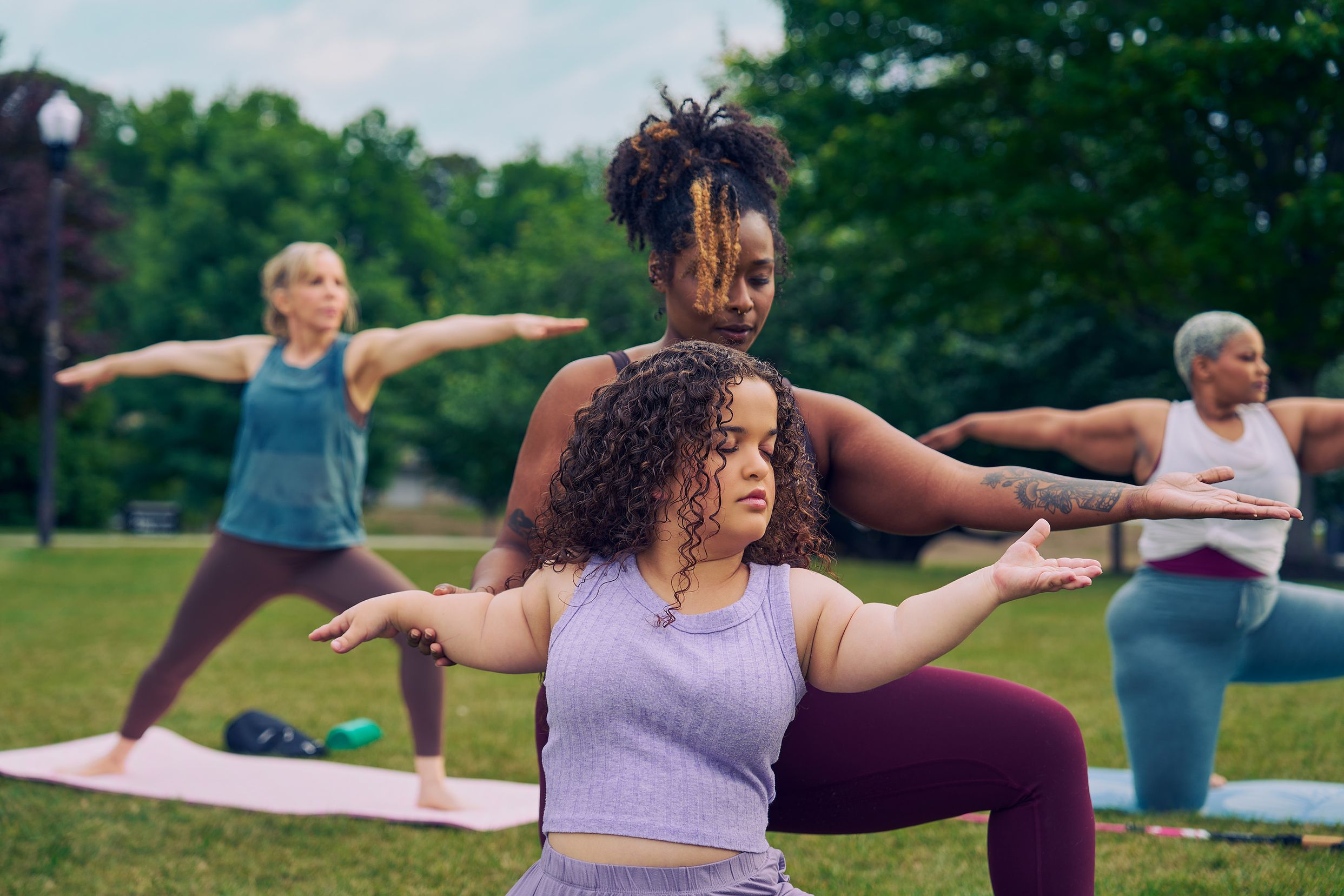 At an outdoor yoga class, a young white woman with dwarfism stands in Warrior pose, with her eyes closed, both arms extended out parallel to the ground, one facing forward and one reaching back. A Black woman yoga instructor kneels behind her and gently guides her arms into the pose. The student has long, curly brown hair and is wearing a purple ribbed tank with comfortable, loose purple pants. The instructor has her black hair pulled back into a natural ponytail with dyed bangs and is wearing a black tank and maroon yoga pants. Behind them, two tall women sit on their mats in the same pose. One of the women in the background is in a modified form of the pose, kneeling. The class is in a grassy park with many large trees.
