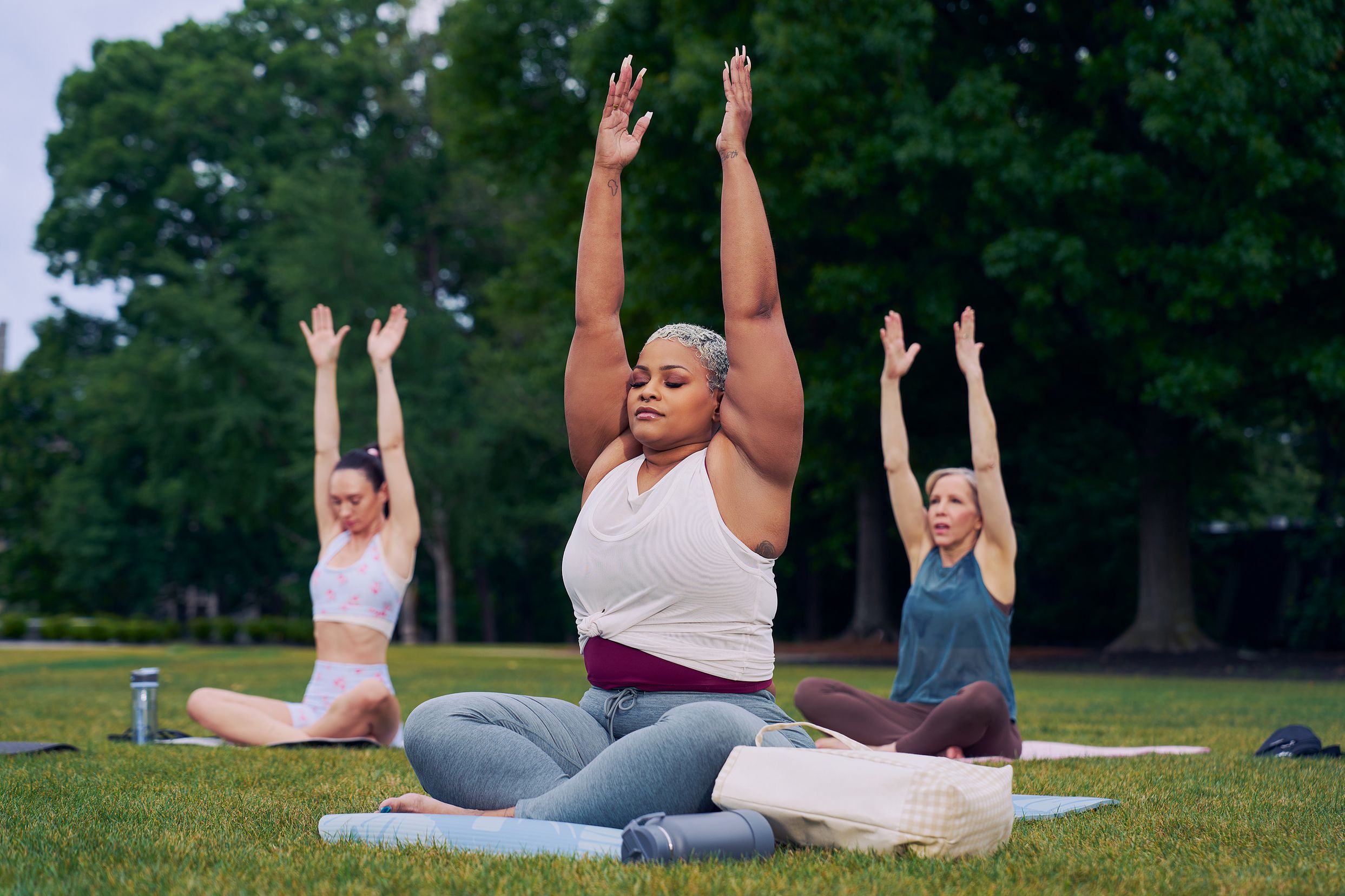 At an outdoor yoga class, a Black woman sits cross-legged with her eyes closed and hands extended straight overhead with palms facing inward toward each other. Her bag and water bottle are next to her. She has icy blonde shortly cut hair and is wearing a white and red layered tank and gray drawstring pants. Behind her, two tall women sit on their mats in the same pose. The class is in a grassy park with many large trees.