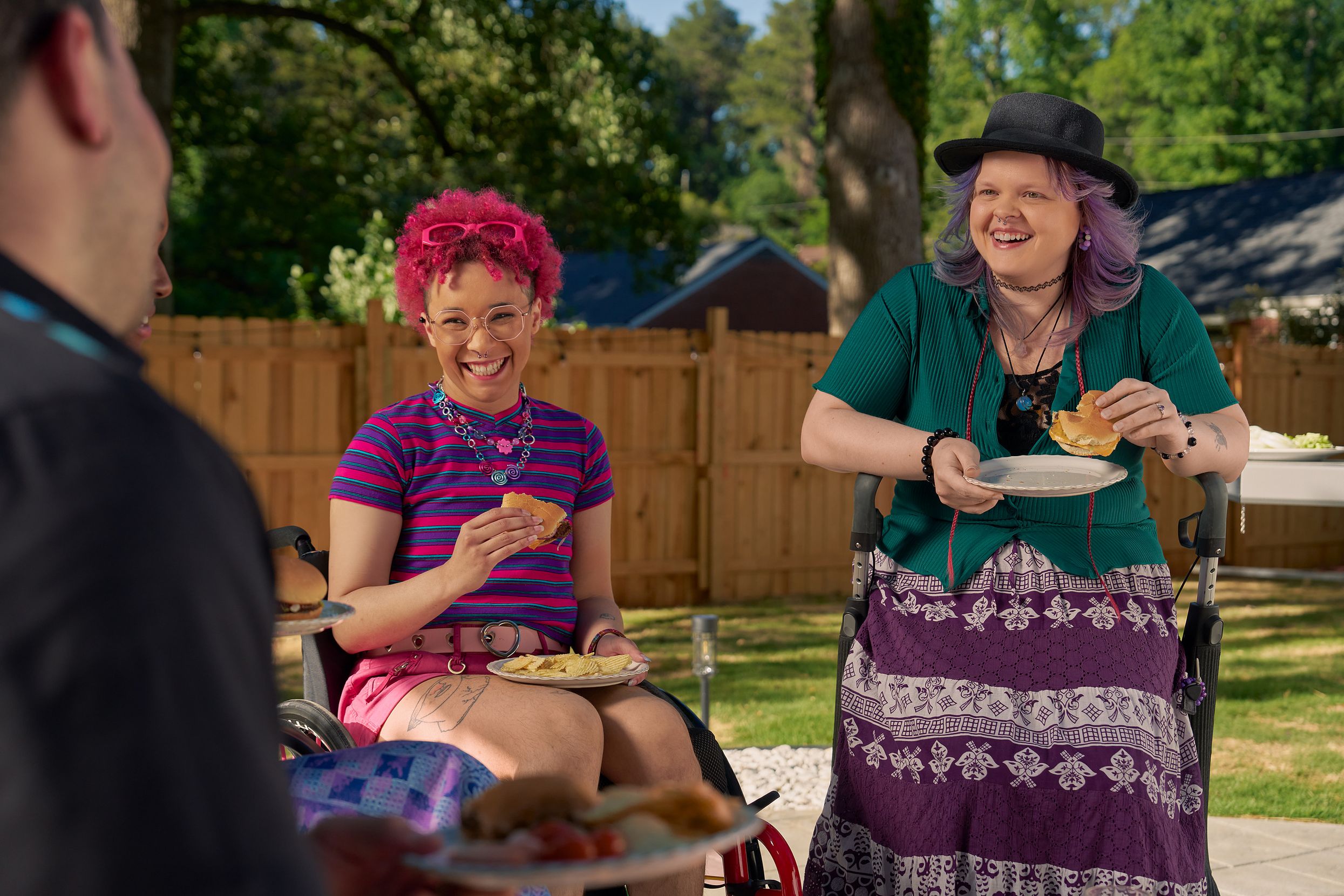 At a backyard barbeque, two nonbinary people sit in their mobility aids, laughing and holding plates of food. One nonbinary person, a Black person with a curly pink fro with loc bangs wearing glasses is seated in a manual wheelchair. They are wearing bright pink sunglassese on their head, a purple and pink striped crop top, pink shorts, and purple sandals. They have several tattoos on both legs. The second nonbinary person, a white person with layered purple hair with two long thin red braids on each side, is seated on a dark gray rollator. They are wearing a black hat, a green shirt, and a purple patterned skirt.
