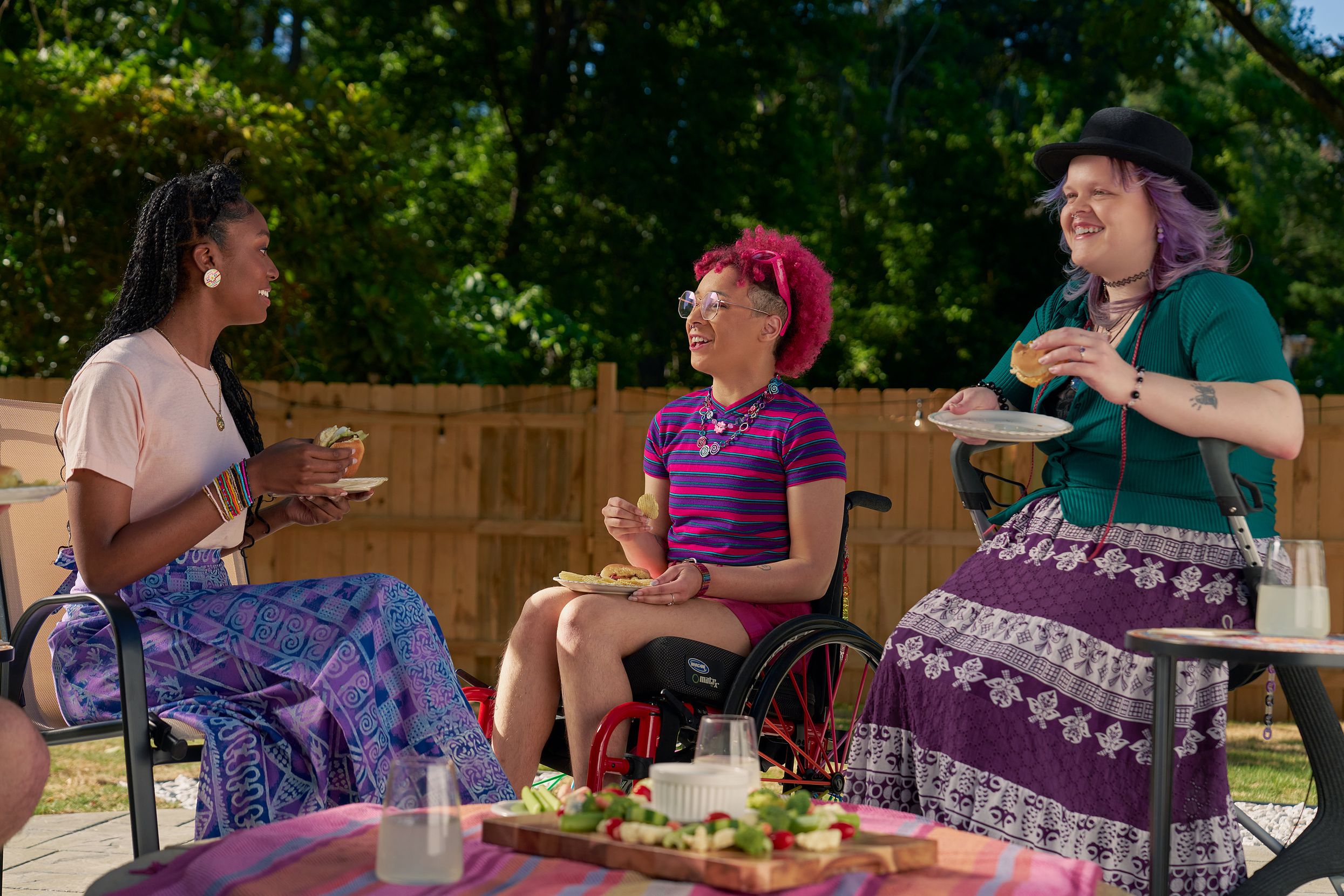 At a backyard barbeque, two nonbinary people sit in their mobility aids, chat with a friend while holding plates of food. One nonbinary person, a Black person with a curly pink fro with loc bangs wearing glasses is seated in a manual wheelchair. They are wearing bright pink sunglassese on their head, a purple and pink striped crop top, pink shorts, and purple sandals. They have several tattoos on both legs. The second nonbinary person, a white person with layered purple hair with two long thin red braids on each side, is seated on a dark gray rollator. They are wearing a black hat, a green shirt, and a purple patterned skirt. Their friend, an African American woman with long braided hair, is wearing a pink shirt and patterned blue and purple skirt.