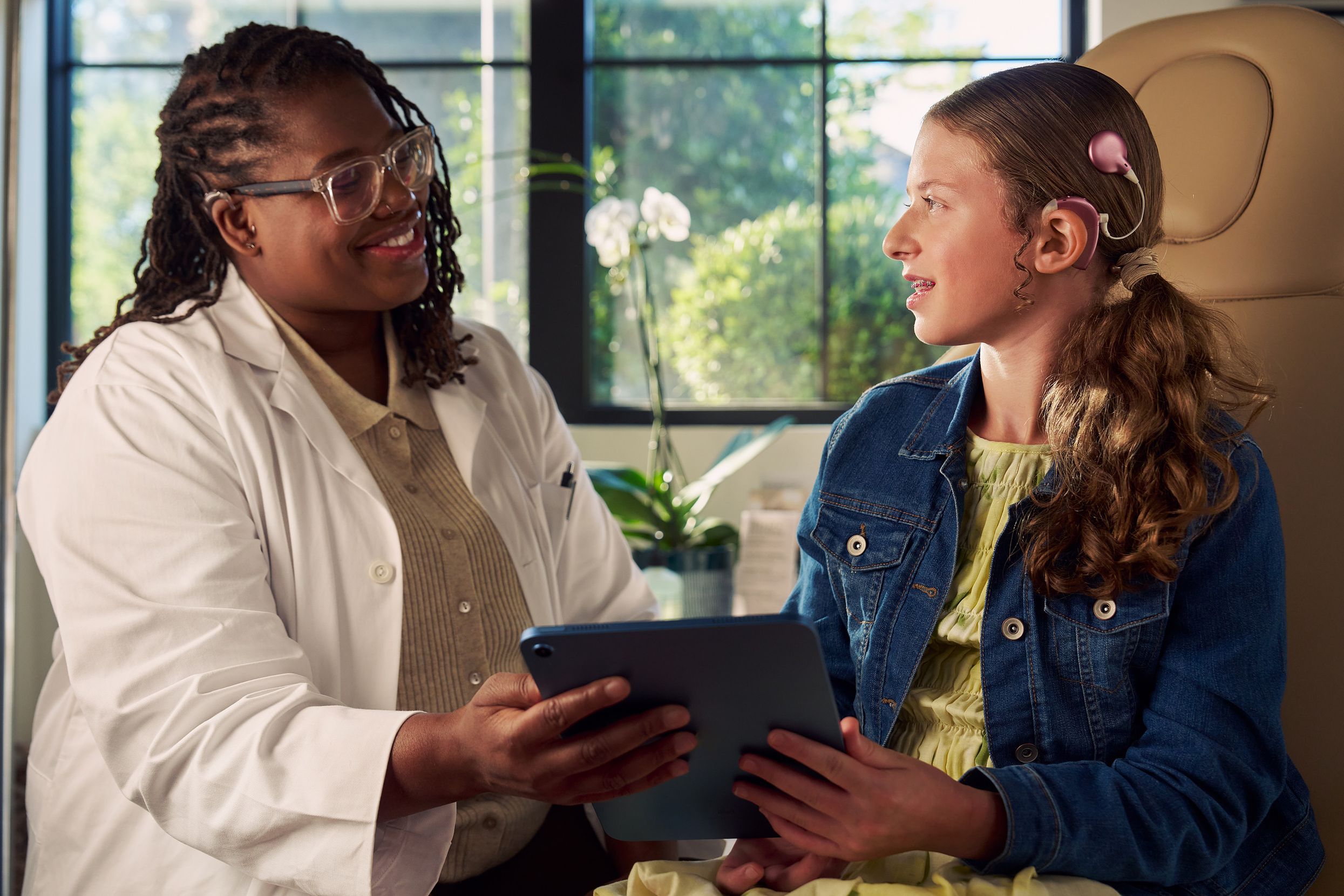 A Black deafBlind audiologist with a cochlear implant and glasses and a young white deaf teen with braces hold a tablet together in a doctor's office. The teen is seated in a medical treatment chair and looks at the audiologist with curiosity. She and has long, wavy sandy blonde hair and is wearing a jean jacket, and a yellow blouse and skirt.