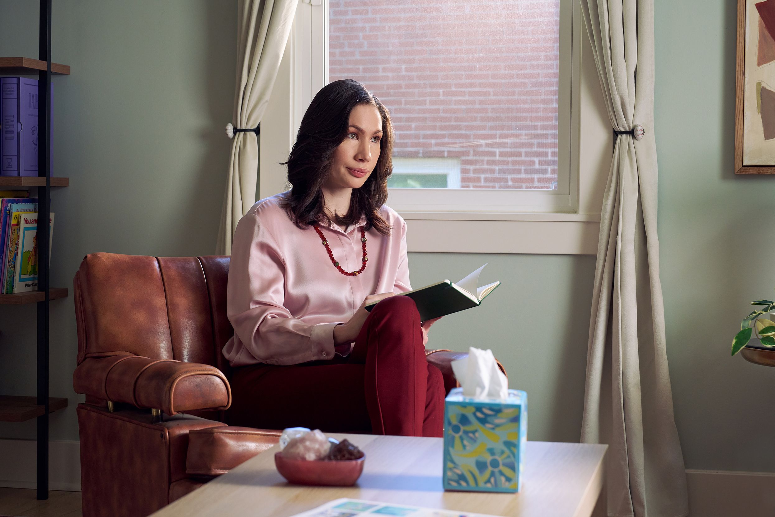 An Eastern European white woman therapist with a facial scar sits in a large chair looks at a client off camera. She is holding an open notebook. A tissue box and a red marble heasrt-shaped bowl of crystals are on a low coffee table in front of her. The therapist's office has several books on display and a window letting in natural light. The therapist is wearing a silk pink top, maroon dress pants, and a minimalist necklace.