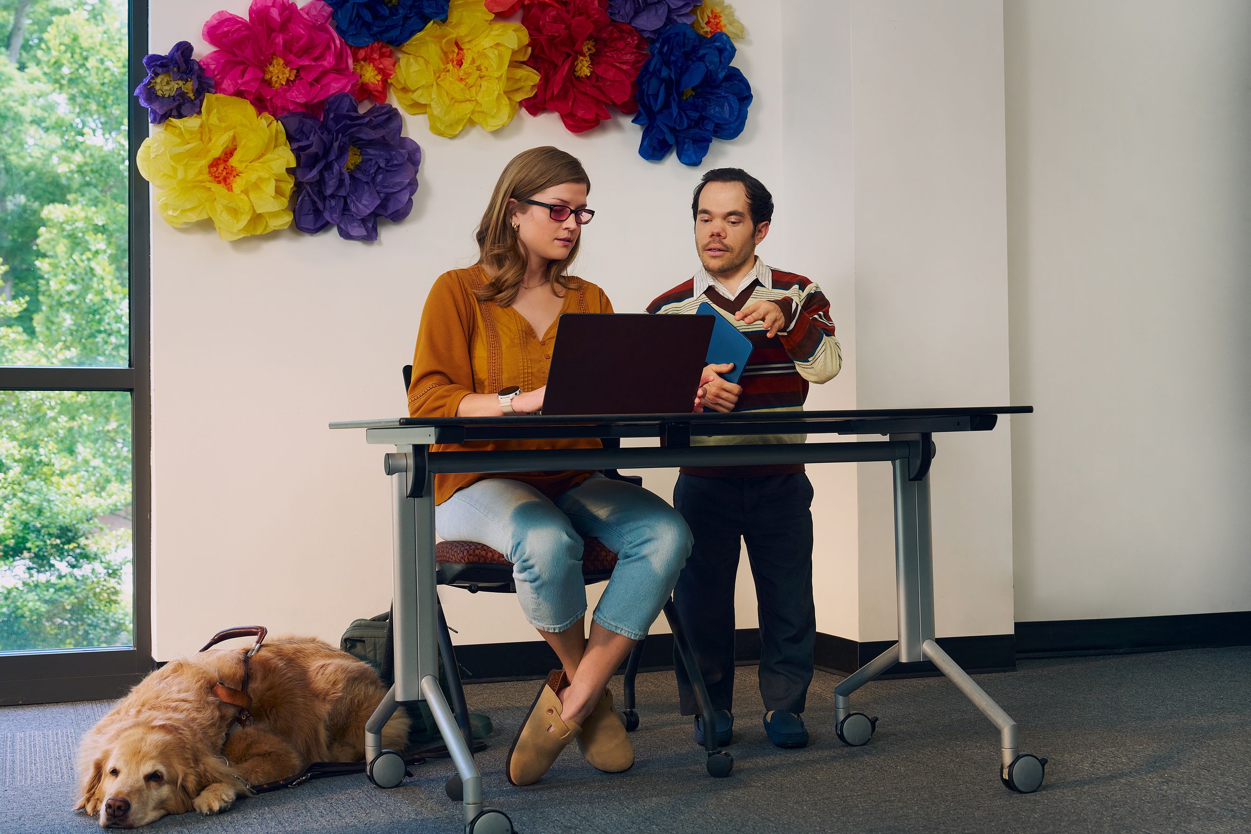 A blind, white woman wearing glasses with red lenses sitting at a lecture table, chats with her professor, a white man with medium brown hair and light brown eyes who has dwarfism, who is standing next to her. They are both looking intently at the student's laptop. A golden retriever service dog wearing a guide harness lays on the floor next to her. The student has dark blonde hair and is wearing an orange blouse and jean capris. A large flower sculpture hangs on the wall behind the lecture table. Next to the sculpture is a large window showing many large trees.