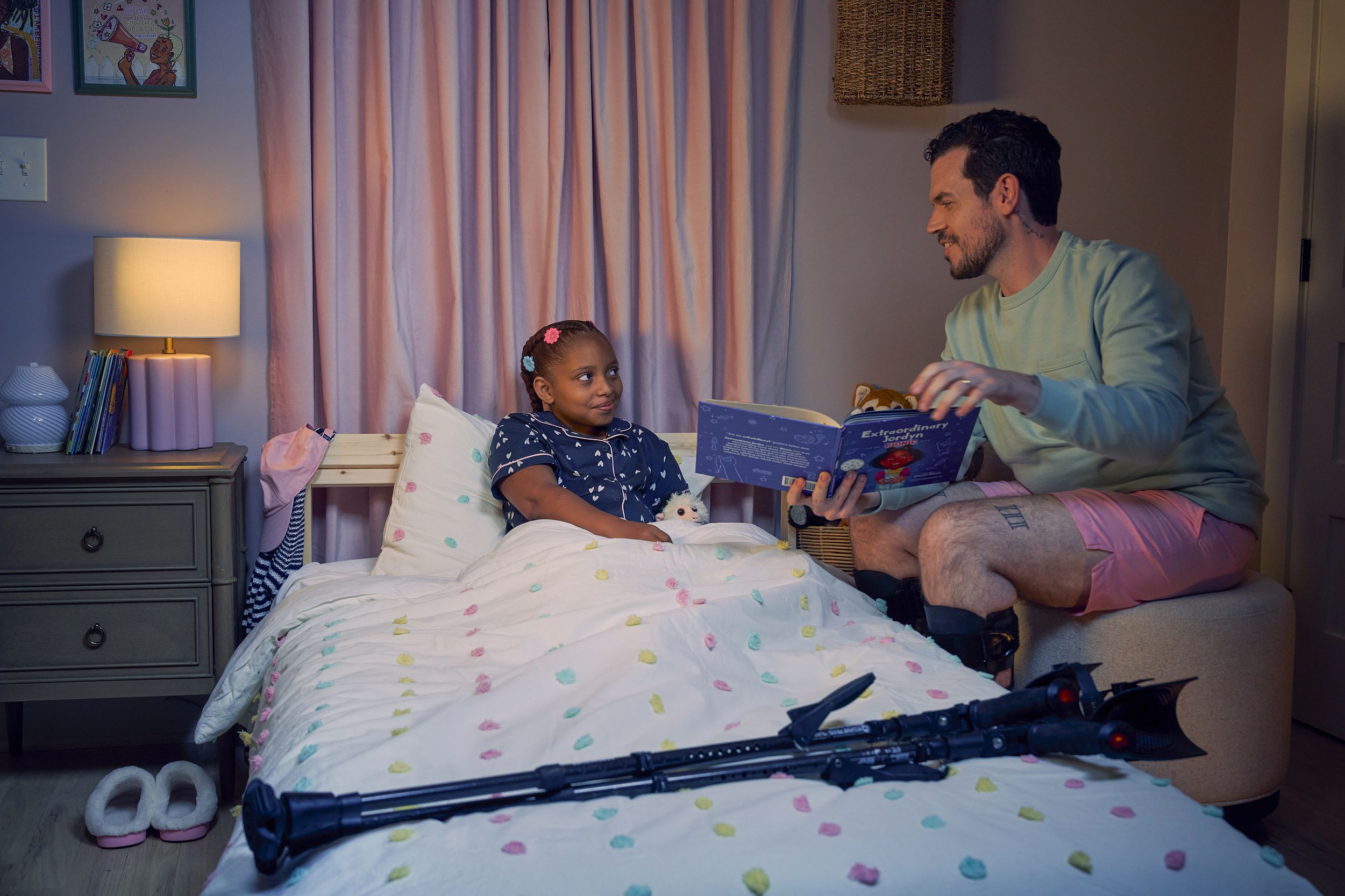 A white man with Cerebral Palsy who wears leg braces holds a children's book titled "Extraordinary Jordyn and Her Bionic Ears" while sitting on an ottoman stool in his daughter's bedroom at night. His daughter is a Black multiracial American girl with thick braided red hair adorned with two flower barrettes. She is tucked into bed and is wearing dark blue patterned pajamas and looks up at her father with a soft smile. The father's black forearm crutches lie on the foot of the bed. He has dark brown curly hair, short brown facial hair, and is wearing a light green sweater with pink shorts. He has a tattoo of the Roman numeral two followed by a slash and the number three. The child's room is painted light pink and includes a nightstand, lamp, a pair of slippers, curtains, and children's books. Her bedspread is white with blue, pink, and yellow polka dots. Framed illustrations of Black women hang on the wall.