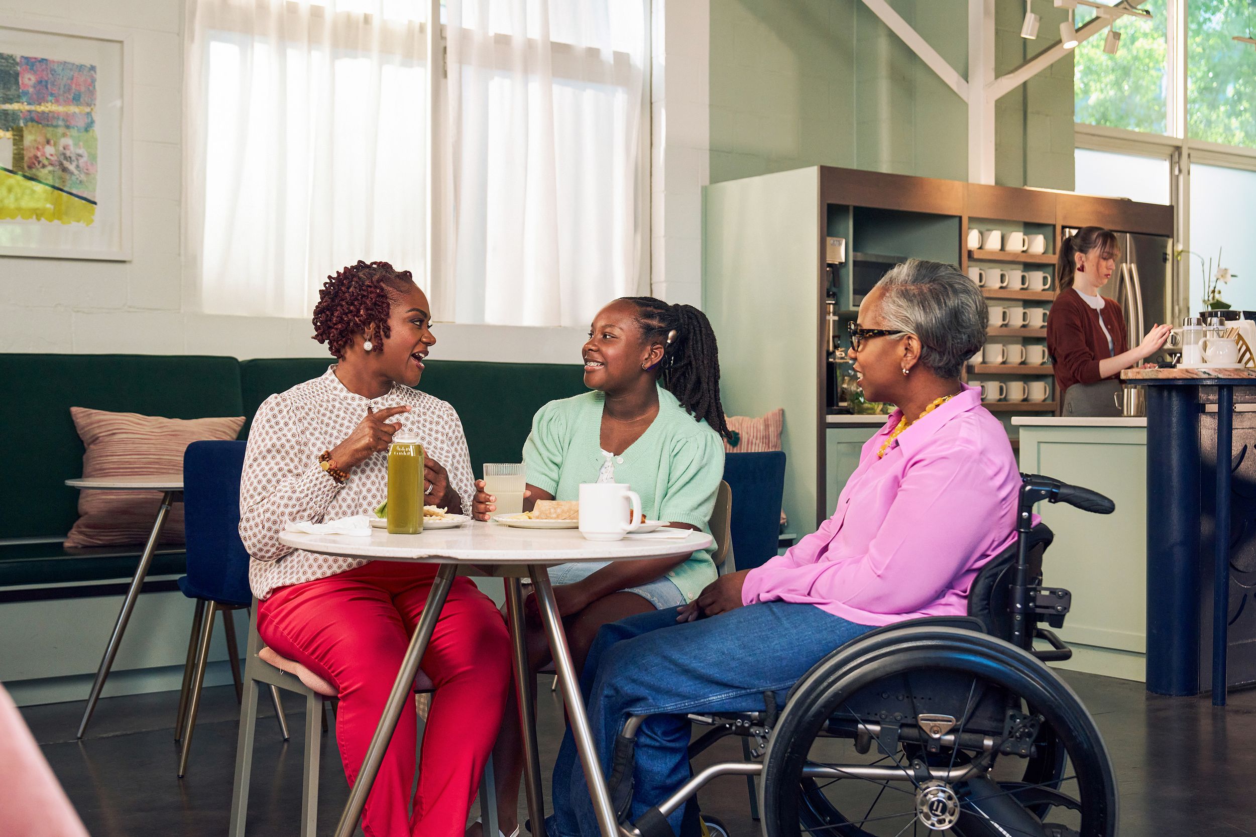 An African American mother and daughter chat at a table in a modern cafe, while the girl's auntie smiles and watches. The auntie is paraplegic and is seated in a manual wheelchair. She has short gray hair and is wearing a lavender silk blouse and dark blue jeans. The mother has short tightly curled red hair and is wearing a patterned blouse and red pants. The daughter is wearing a cochlear implant and has her braided hair pulled back into a pony tail. She is wearing a light green cardigan and jean shorts. Behind them, a barista prepares drinks.