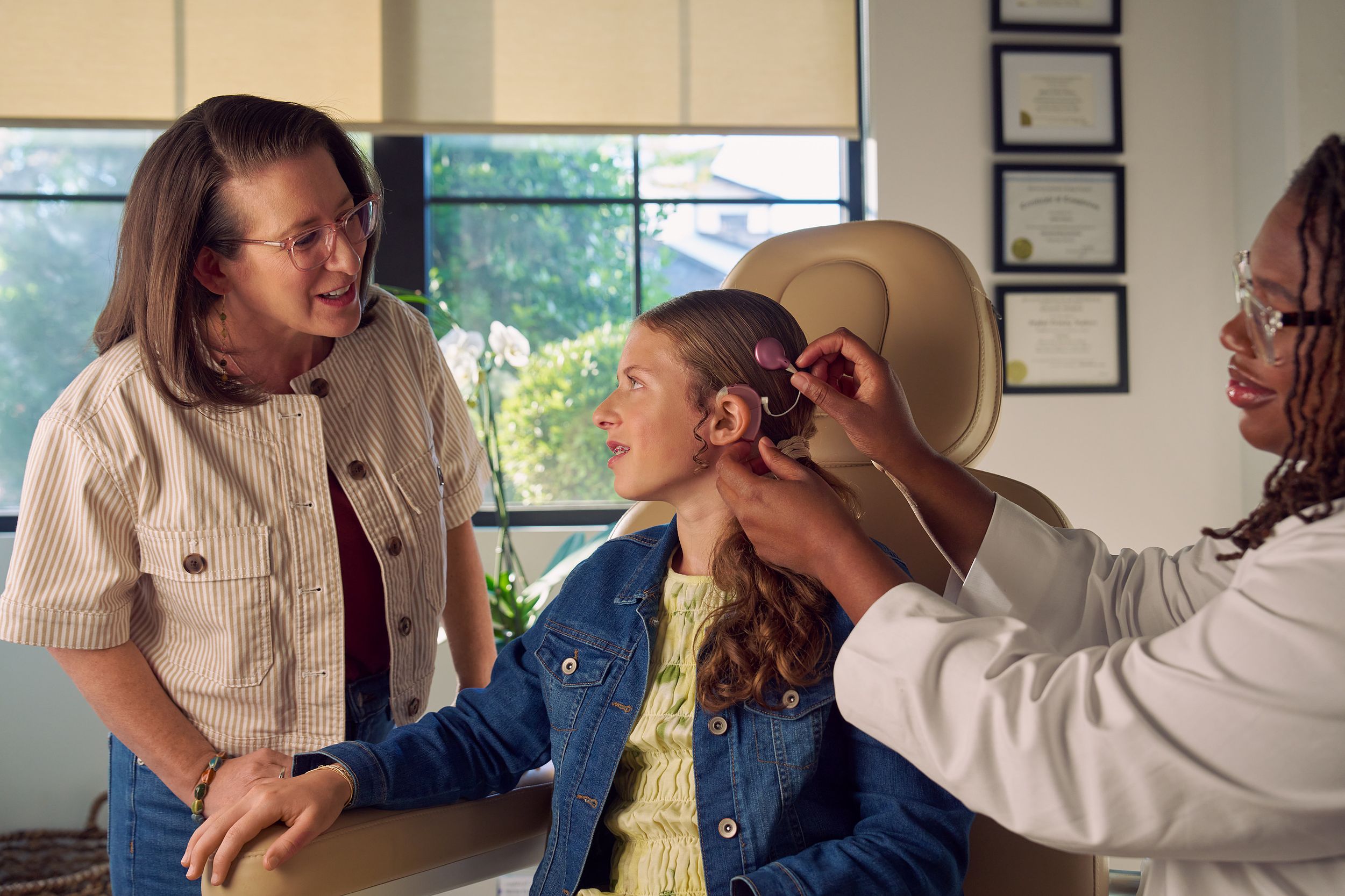 A young white deaf teen with braces sits in a medical treatment chair while an audiologist places a mauve colored cochlear implant on her head. The teen looks at her mother, who is standing and looking at the procedure with curiosity. The teen has long, wavy sandy blonde hair and is wearing a jean jacket, and a yellow blouse and skirt. The audiologist is a Black deafBlind woman wearing glasses and has dark brown shoulder length locs. The teen's mother is a middle-aged white woman with straight brown hair and glasses. She is wearing an off-white striped summer jacket and jeans. A row of four framed professional degrees are hanging on the wall behind them.
