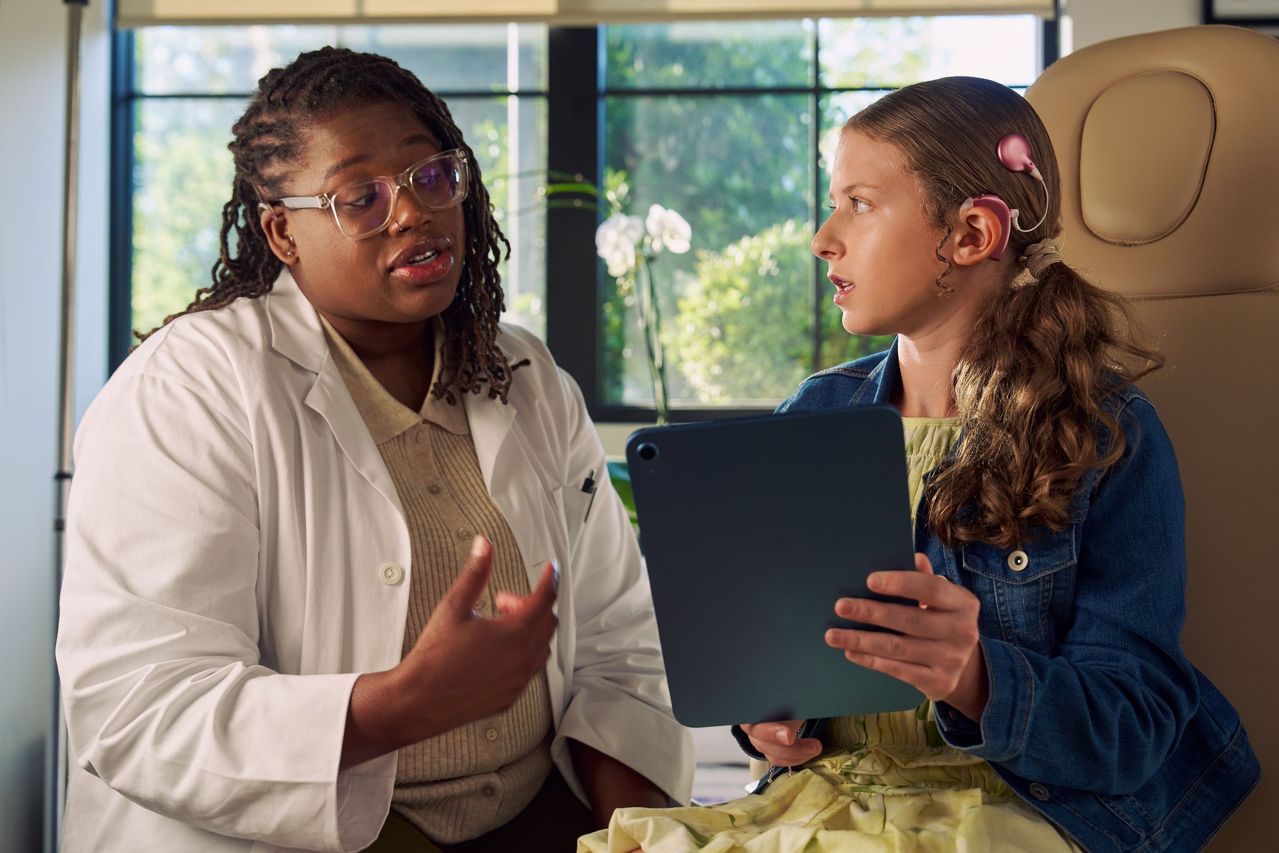 A Black deafBlind audiologist with a cochlear implant and glasses talks with a young white deaf teen with braces holding a tablet. The teen is seated in a medical treatment chair and looks at the audiologist with curiosity. She and has long, wavy sandy blonde hair and is wearing a jean jacket, and a yellow blouse and skirt.