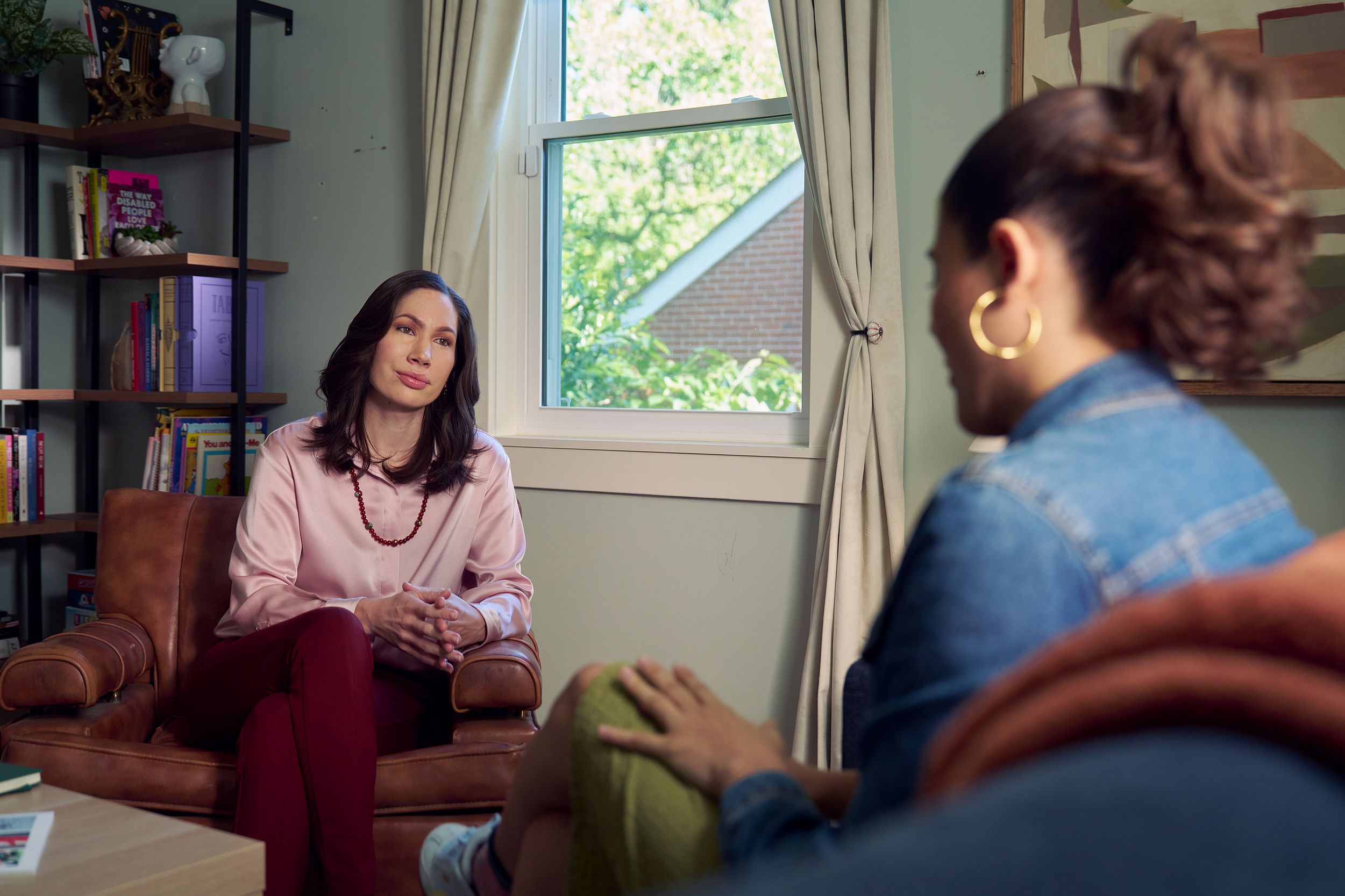 An Eastern European white woman therapist with a facial scar sits in a large chair and looks at a client seated on a couch in front of her. The client is a Latina woman with dark brown wavy hair pulled into a ponytail. The therapist's office has several books on display, an abstract art piece, and a window letting in natural light. The therapist is wearing a silk pink top, maroon dress pants, and a minimalist necklace. The client is wearing gold hoop earrings, a jean jacket, green skirt, and sneakers.