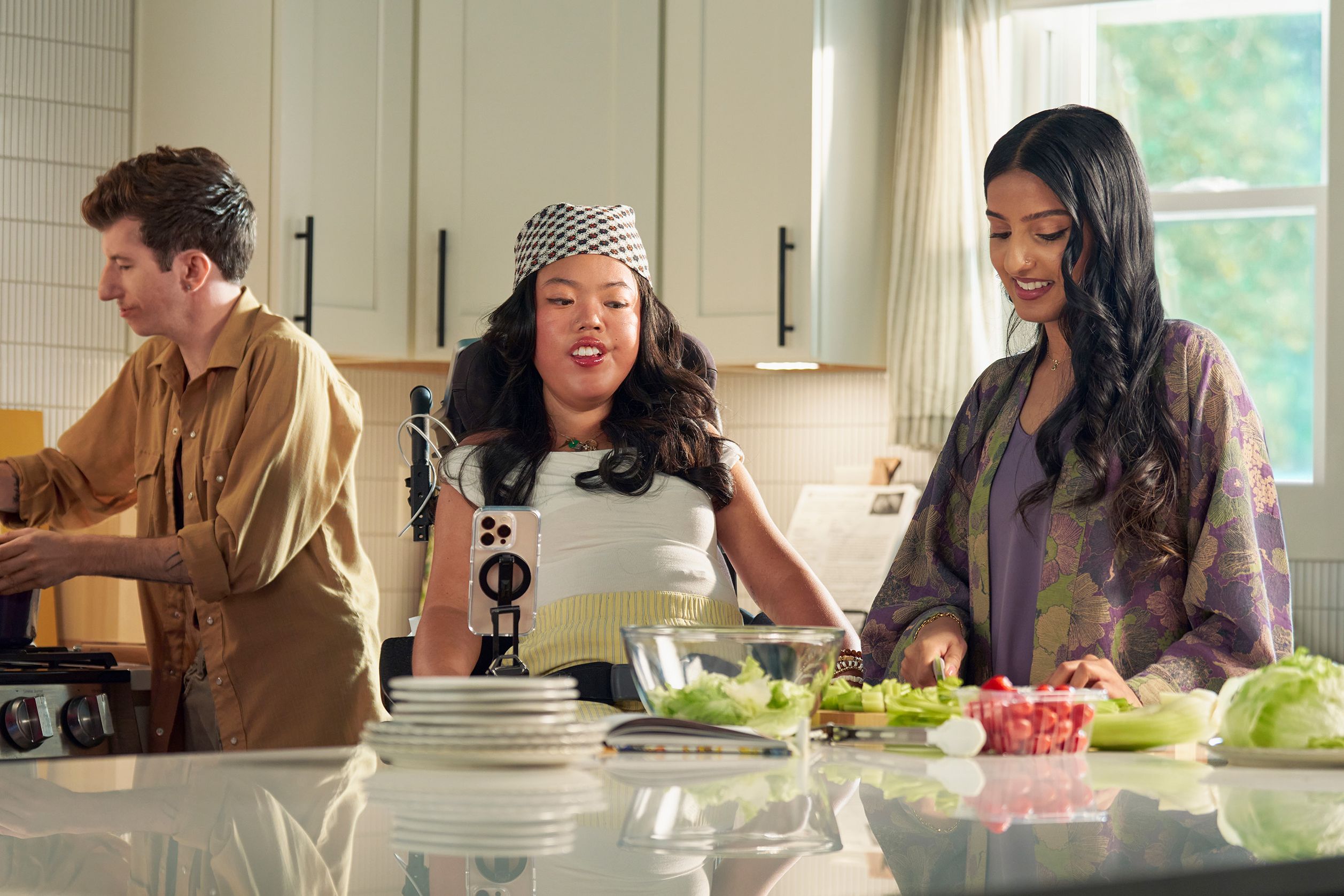 A SouthEast Asian woman with curled black hair seated in her power wheelchair watches her roommate prepare a salad in their bright kitchen. Her smart phone is mounted to her wheelchair. Behind them, a white male with Moebius Syndrome stirs a pot on the stove. The SouthEast Asian woman is wearing a white top with striped, yellow linen pants and a checkered headscarf. Her roommate has long dark hair and is wearing a purple floral dress.