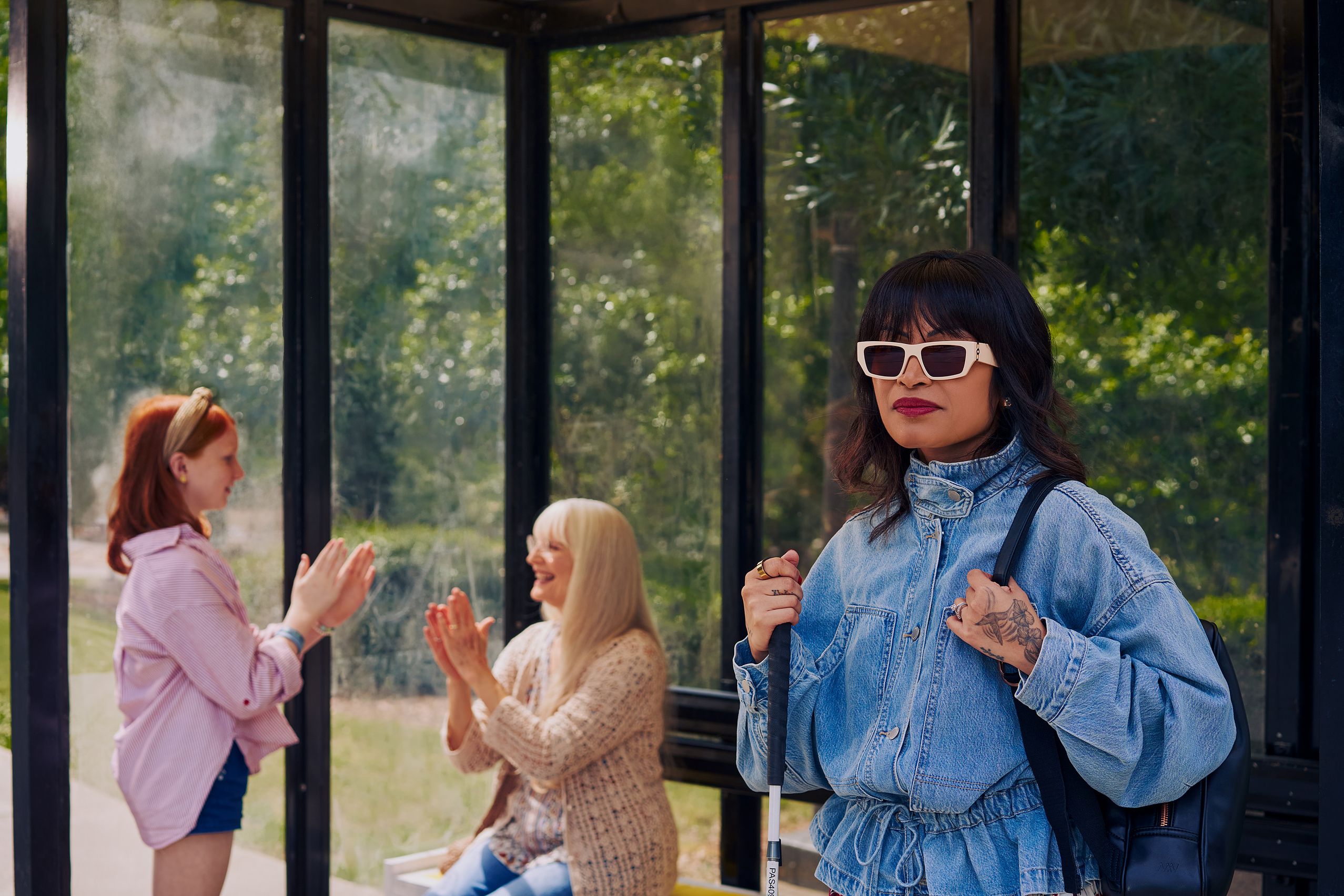 An Asian Filipino woman with Retinitis Pigmentosa and Usher Syndrome stands at a covered bus stop. She is wearing white sunglasses and is holding her white cane with her tattooed hands. She has on a jean jacket and a black backpack. Behind her, a white European girl with red hair plays pattycake hand game with her grandmother, who is sitting on the bus stop bench. Behind the bus stop are several large trees and walking paths.