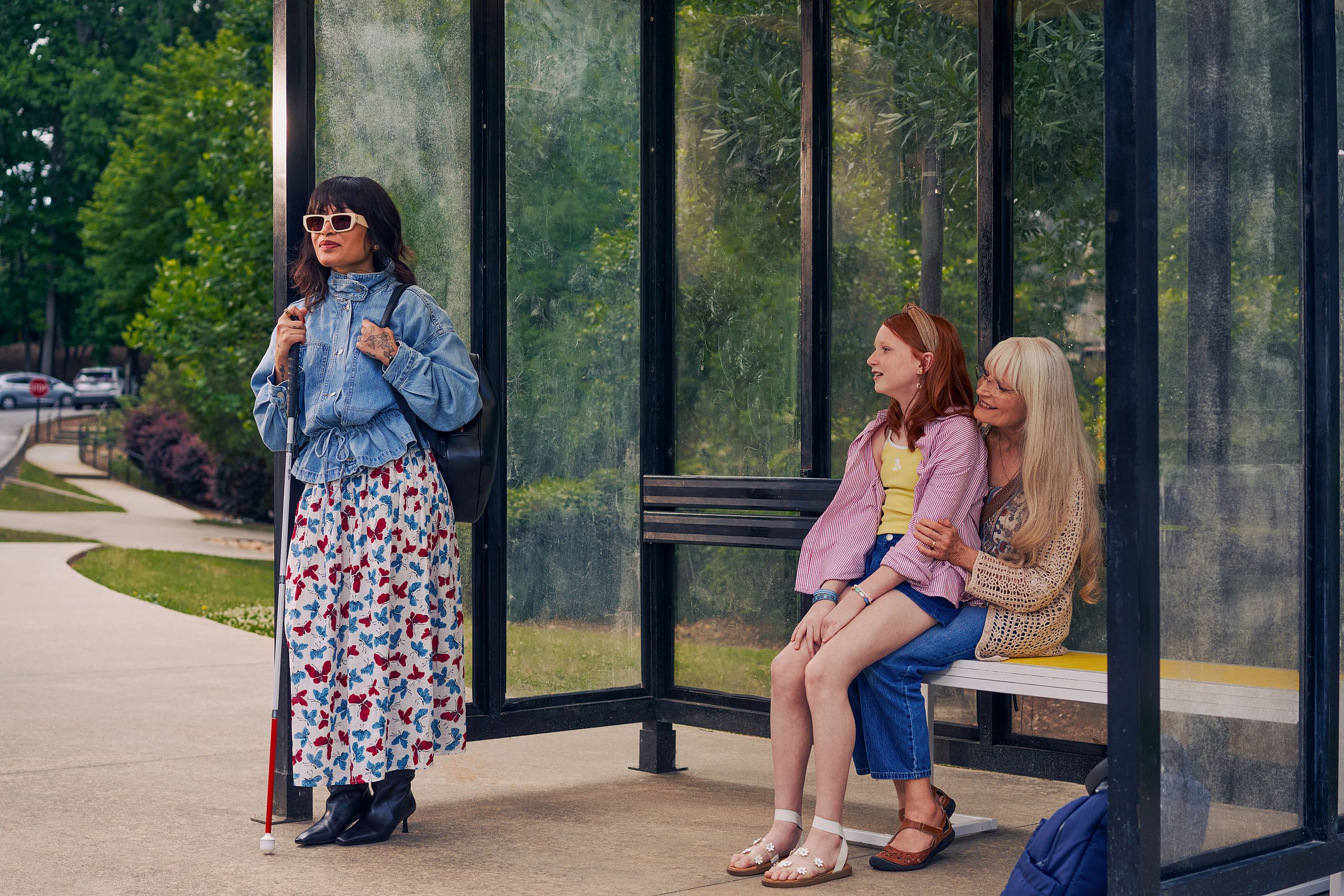 An Asian Filipino woman with Retinitis Pigmentosa and Usher Syndrome stands at a covered bus stop. She is wearing white sunglasses and is holding her white cane with her tattooed hands. She has on a jean jacket, a butterfly patterned skirt, and a black backpack. Behind her, a white European girl with red hair sits on her grandmother's lap on the bus stop bench. Behind the bus stop are several large trees and walking paths.