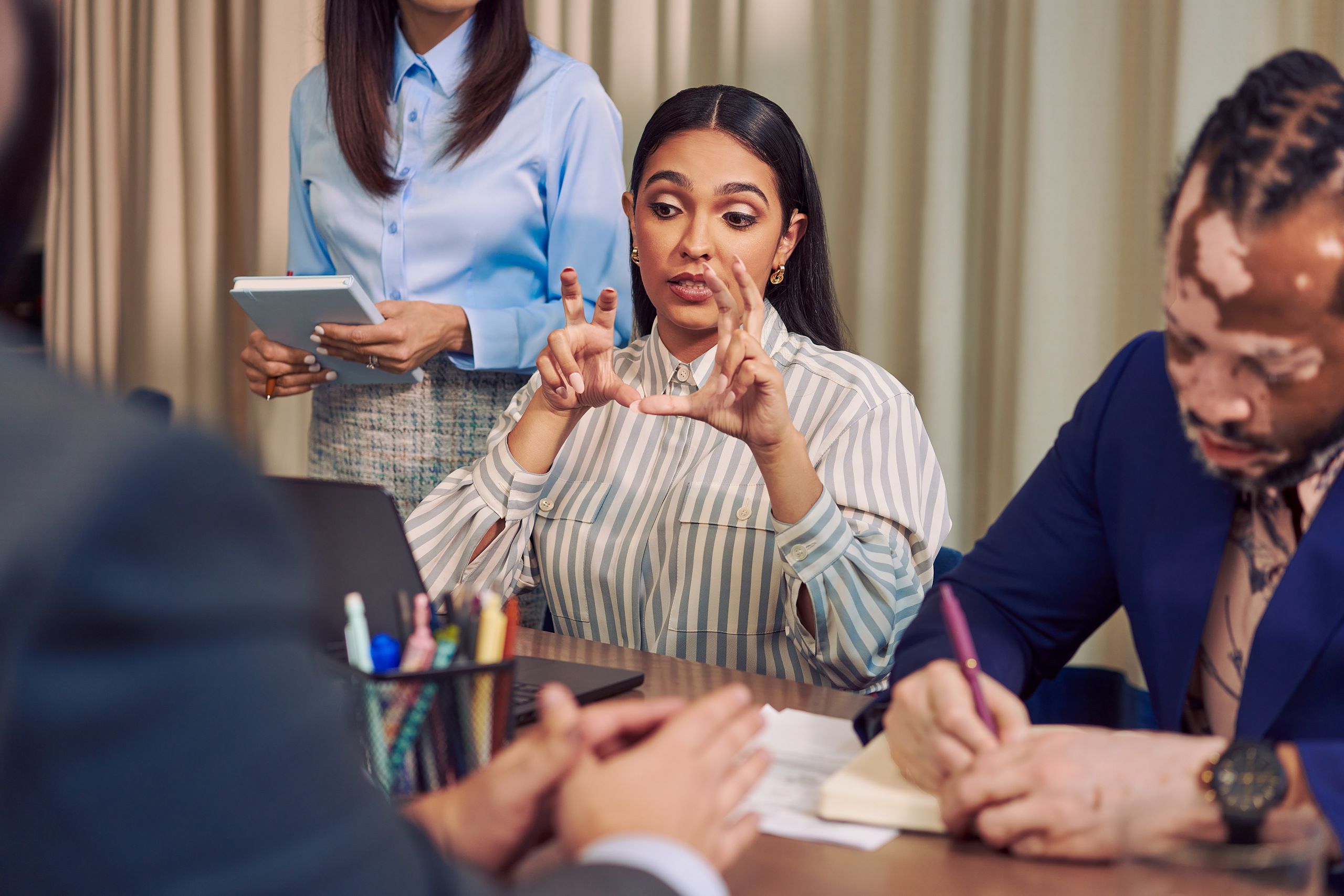 A Deaf Latina Puerto Rican woman uses American Sign Language while seated at a conference room table with three colleagues. She has long deep brown hair and is wearing a striped business shirt and hoop earrings. One colleague, an African American man with olive and porcelain skin, Non-Segmental Vitiligo, and brown barrel-twist dreadlocks writes in a notebook next to her. He is wearing a blue business suit.