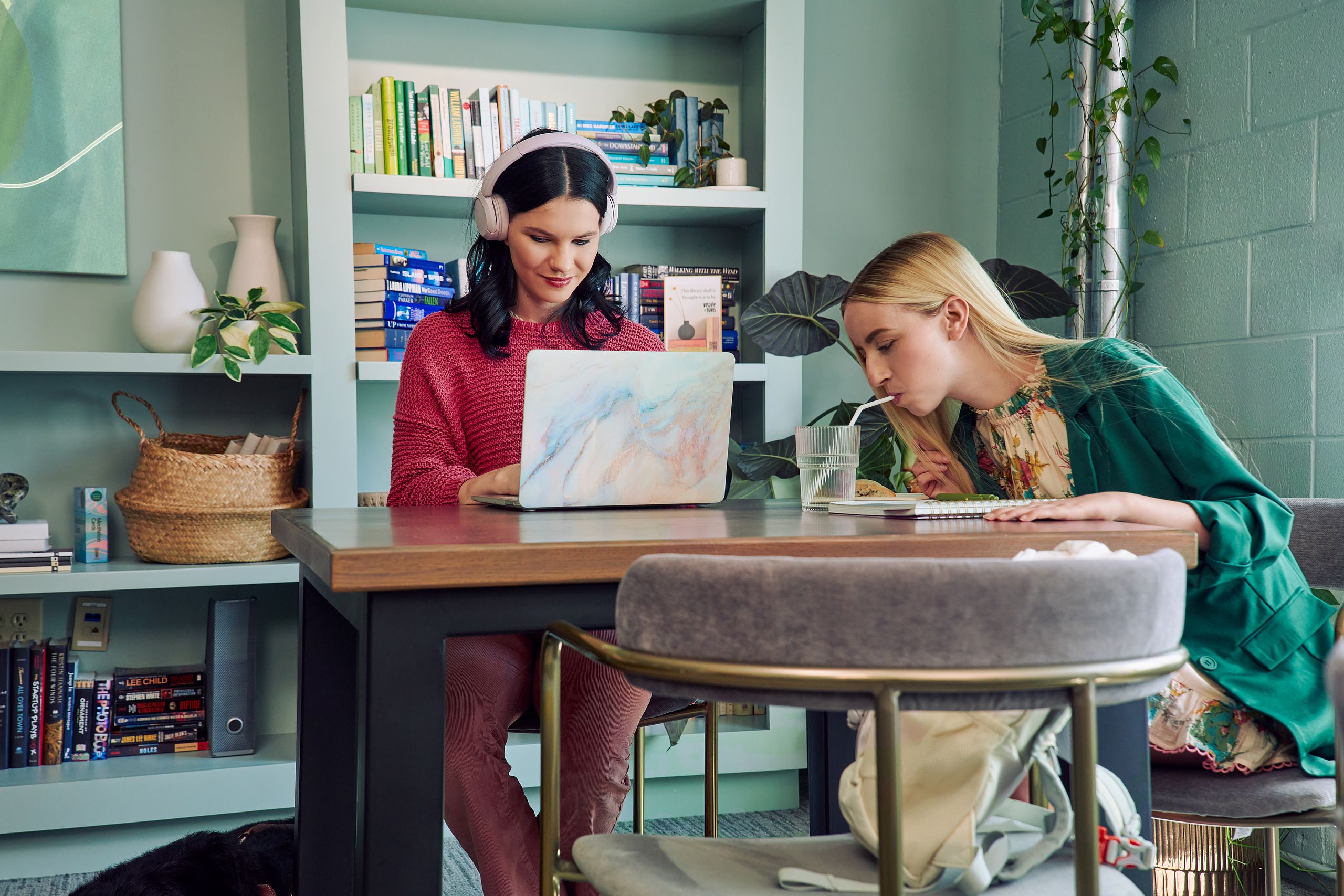 A young white woman with long, straight blonde hair and partially paralyzed arms and hands bends forward to drink water from a cup through a straw. She is sitting at a table in a modern coworking office space with a young pale white blind woman, who is typing on a laptop computer. The blind woman has shiny black hair styled in waves and is wearing over the ear headphones, a chunky knitted pink sweater, and dusky pink flared pants. The woman with partially paralyzed arms and hands is wearing a floral dress with pink trim and a green blazer. The coworking office space is painted a warm mint green and has a large bookcase and features several books, plants, a rattan basket, and clay vases.