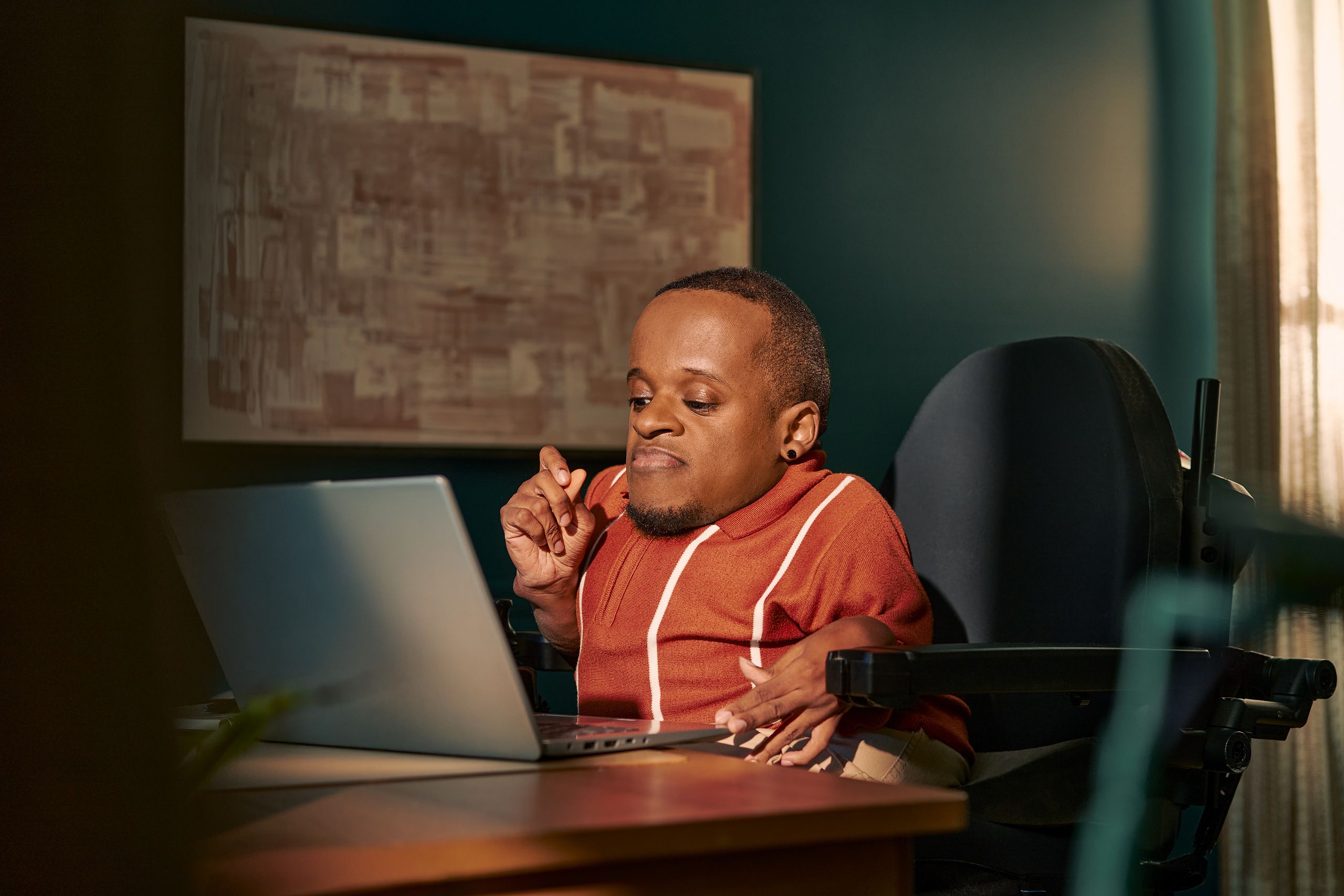A black man who is a little person is seated in his power wheelchair in a dark teal home office. He types on a laptop on his desk, his hand held up in concentration. An abstract art piece is on the wall behind him. He has closely cropped black hair, brown eyes, and is wearing a rust-orange knit quarter-zip sweater with white vertical stripes, light khaki trousers, and a small stud earring.
