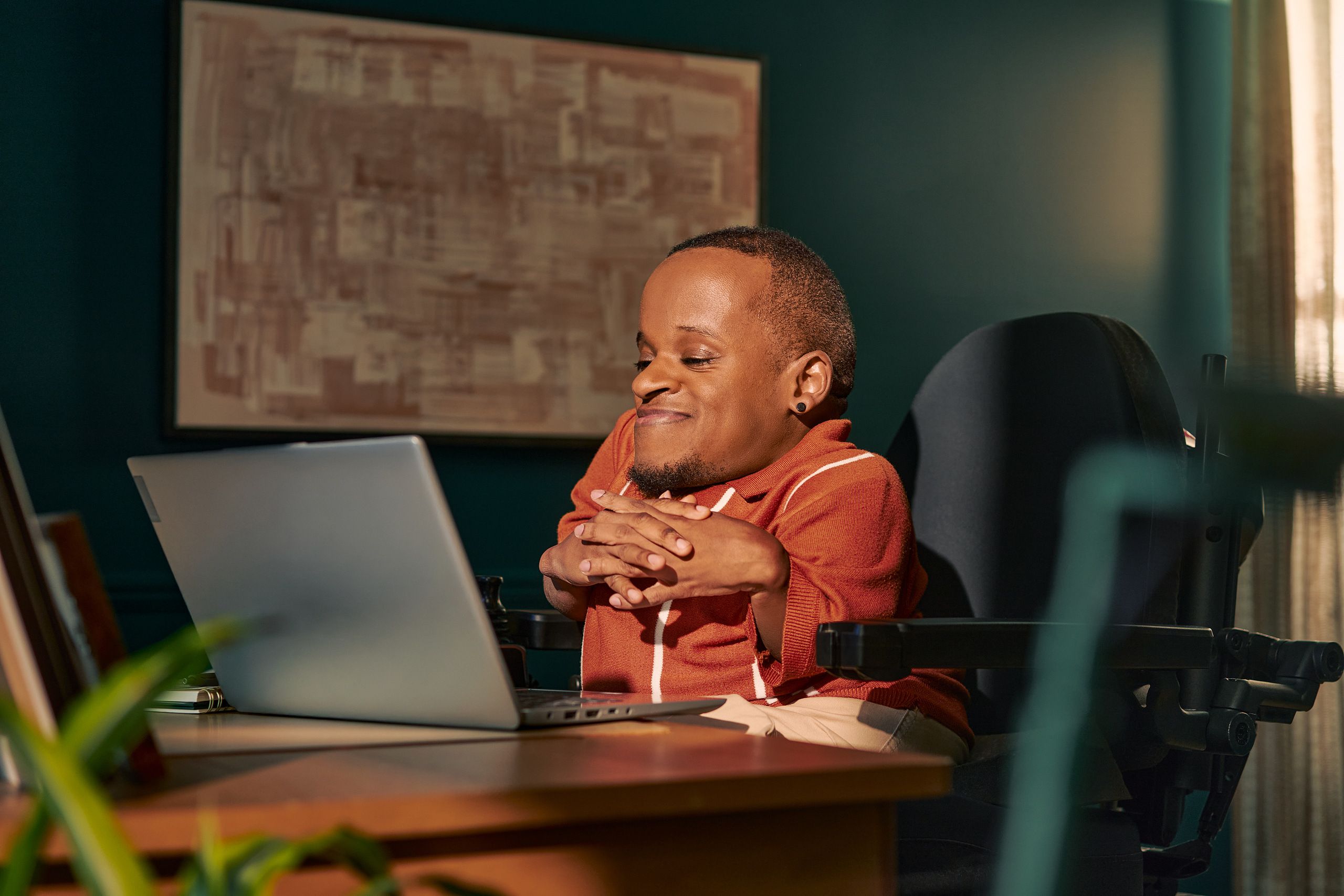 A black man who is a little person is seated in his power wheelchair in a dark teal home office. He smiles down at his open laptop, his hands together with fingers interlaced. A notebook and framed photos are on the desk in front of him. An abstract art piece is on the wall behind him. He has closely cropped black hair, brown eyes, and is wearing a rust-orange knit quarter-zip sweater with white vertical stripes, light khaki trousers, and a small stud earring.