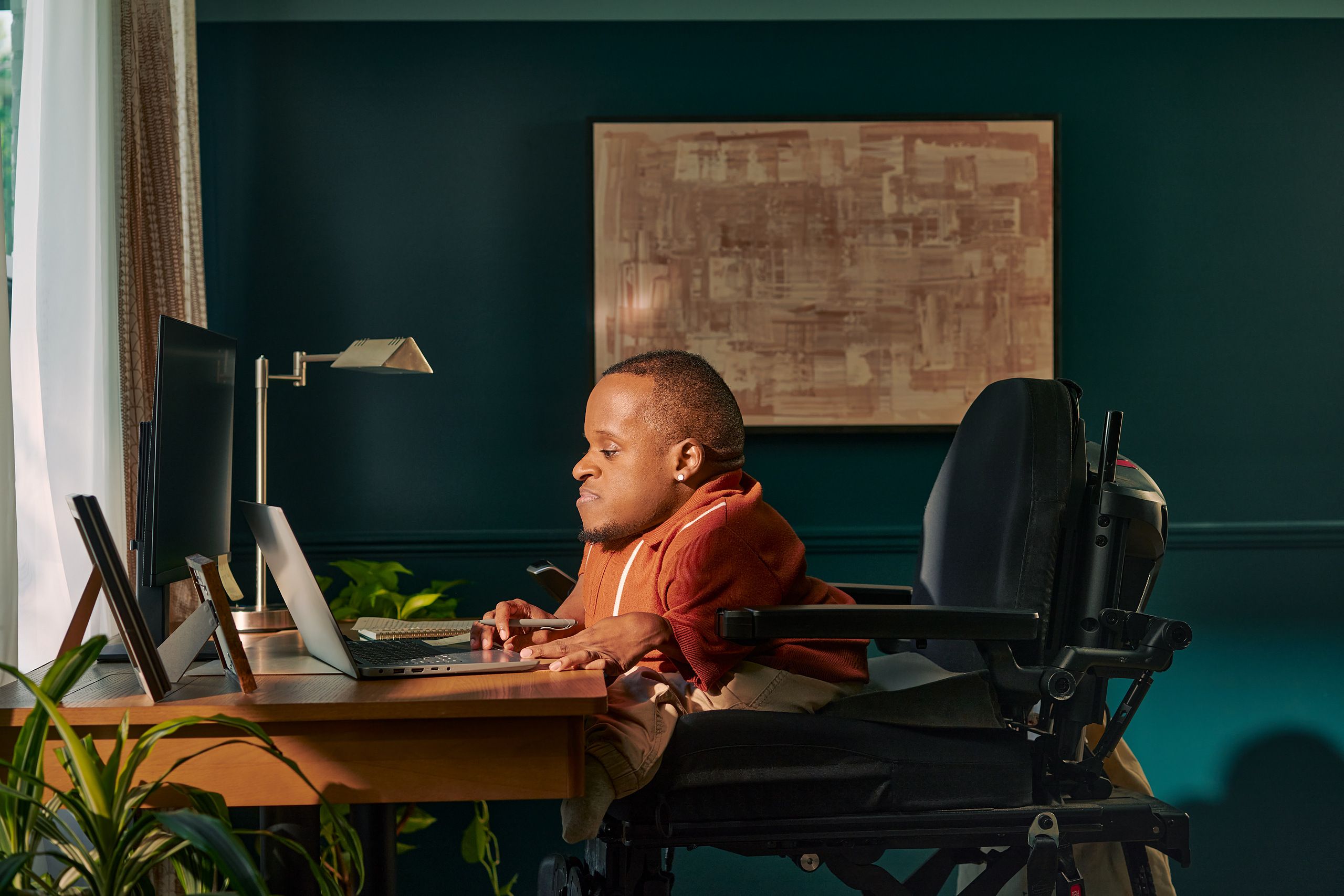 A black man who is a little person is seated in his power wheelchair in a dark teal home office. He holds a pen and types on his laptop. A monitor, notebook, pen, lamp, and framed photos are on the desk in front of him. His wheelchair is in an elevated position, allowing him to be level with his desk. He is facing a window, letting in light through white and patterned curtains. His desk is surrounded by several house plants and an abstract art piece. He has closely cropped black hair, brown eyes, and is wearing a rust-orange knit quarter-zip sweater with white vertical stripes, light khaki trousers, and a small stud earring. A tote bag hangs on the back of his wheelchair.