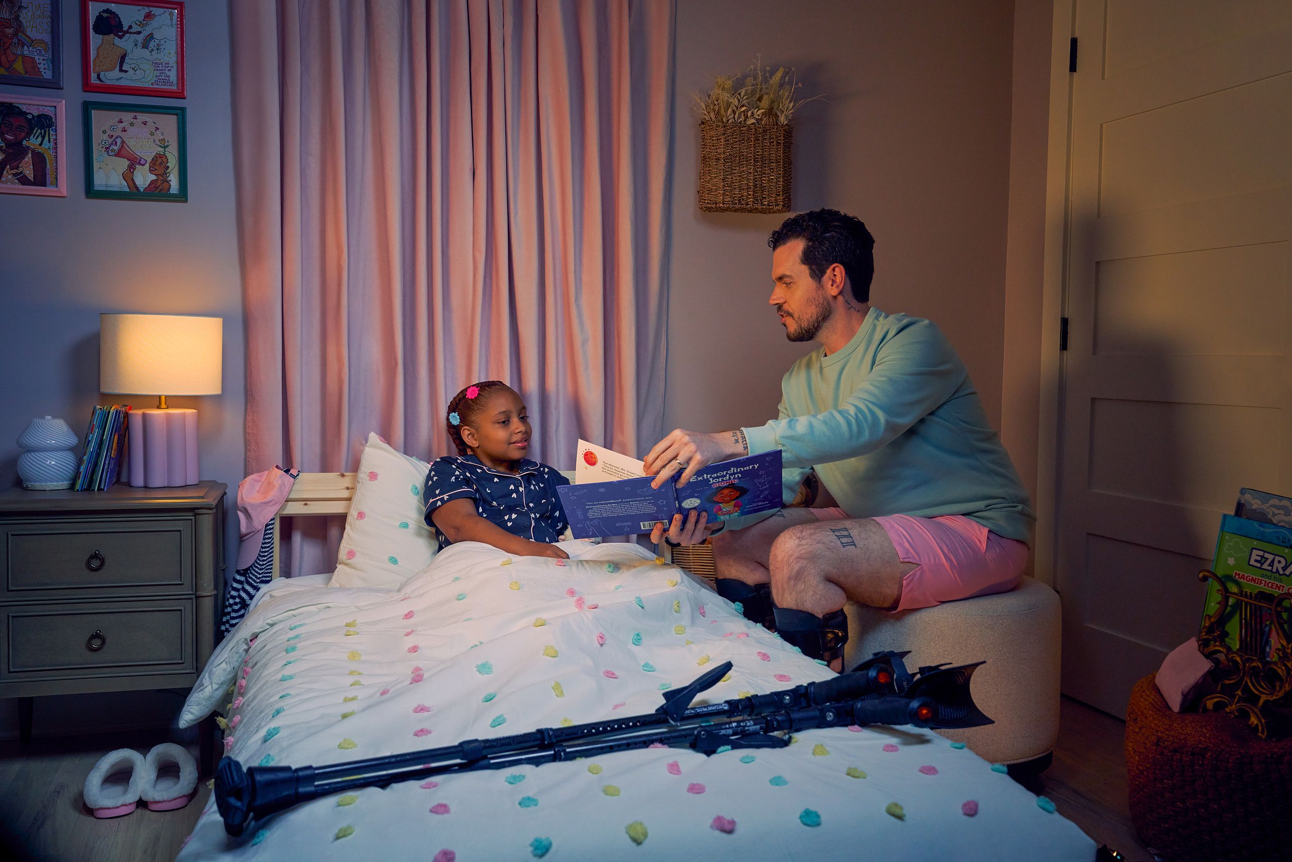 A white man with Cerebral Palsy who wears leg braces shows a children's book titled "Extraordinary Jordyn and Her Bionic Ears" to his daughter in her bedroom at night. His daughter is a Black multiracial American girl with thick braided red hair adorned with two flower barrettes. She is tucked into bed and is wearing dark blue patterned pajamas and looks at the open book. The father's black forearm crutches lie on the foot of the bed. He has dark brown curly hair, short brown facial hair, and is wearing a light green sweater with pink shorts. He has a tattoo of the Roman numeral two followed by a slash and the number three. The child's room is painted light pink and includes a nightstand, lamp, a pair of slippers, curtains, and children's books. Her bedspread is white with blue, pink, and yellow polka dots. Framed illustrations of Black women hang on the wall.