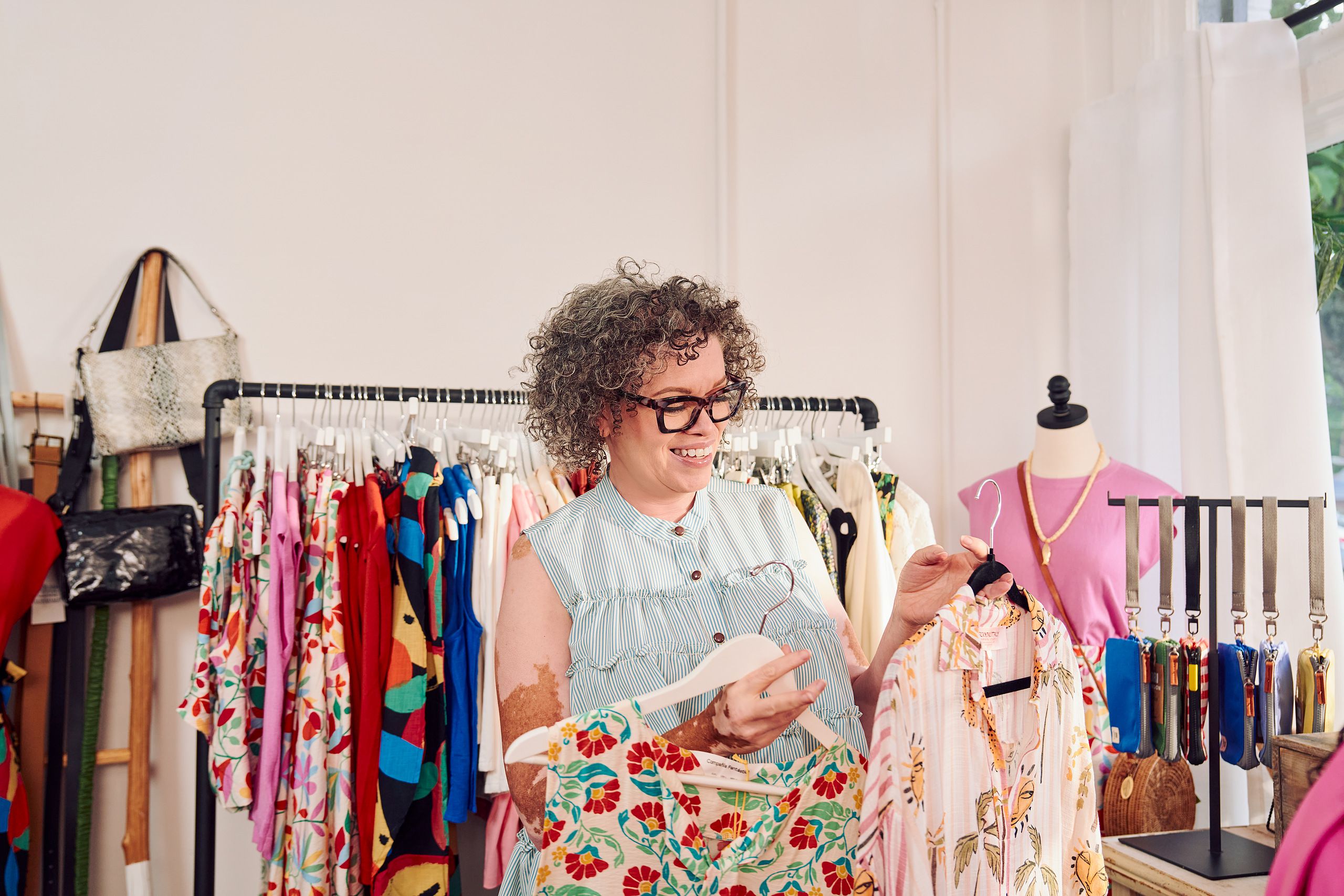 A Black woman shopkeeper with Vitiligo smiles and holds a patterned blouse in each hand, looking down at both options. She has a short, curly bob and is wearing glasses, a striped flowy blouse. Behind her are several clothing racks with high end garments, purses, belts, and mannequins.