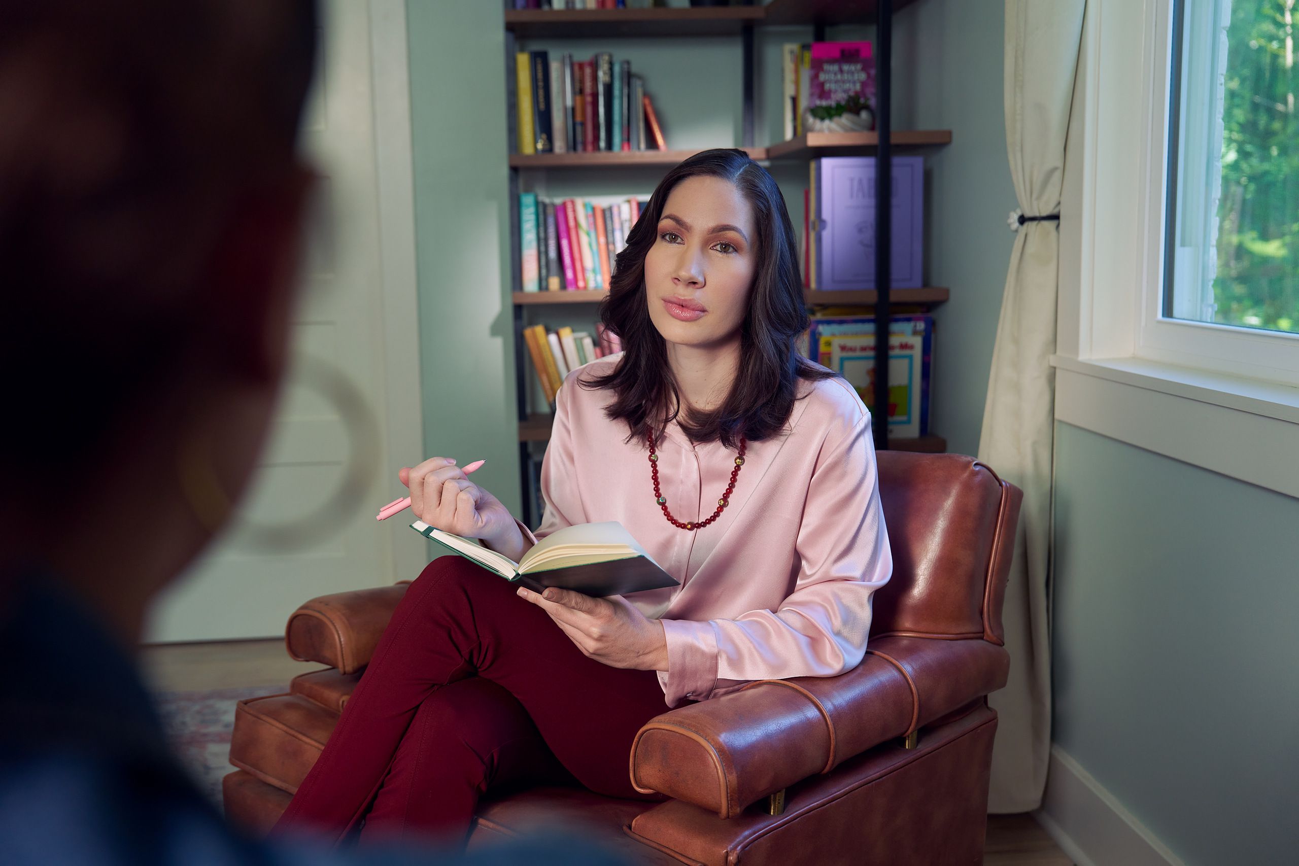 An Eastern European white woman therapist with a facial scar sits in a large chair with an understanding look on her face. She is holding a pen and open notebook, and is looking at a client off camera. The therapist's office has several books on display and a window letting in natural light. The therapist is wearing a silk pink top, maroon dress pants, and a minimalist necklace.