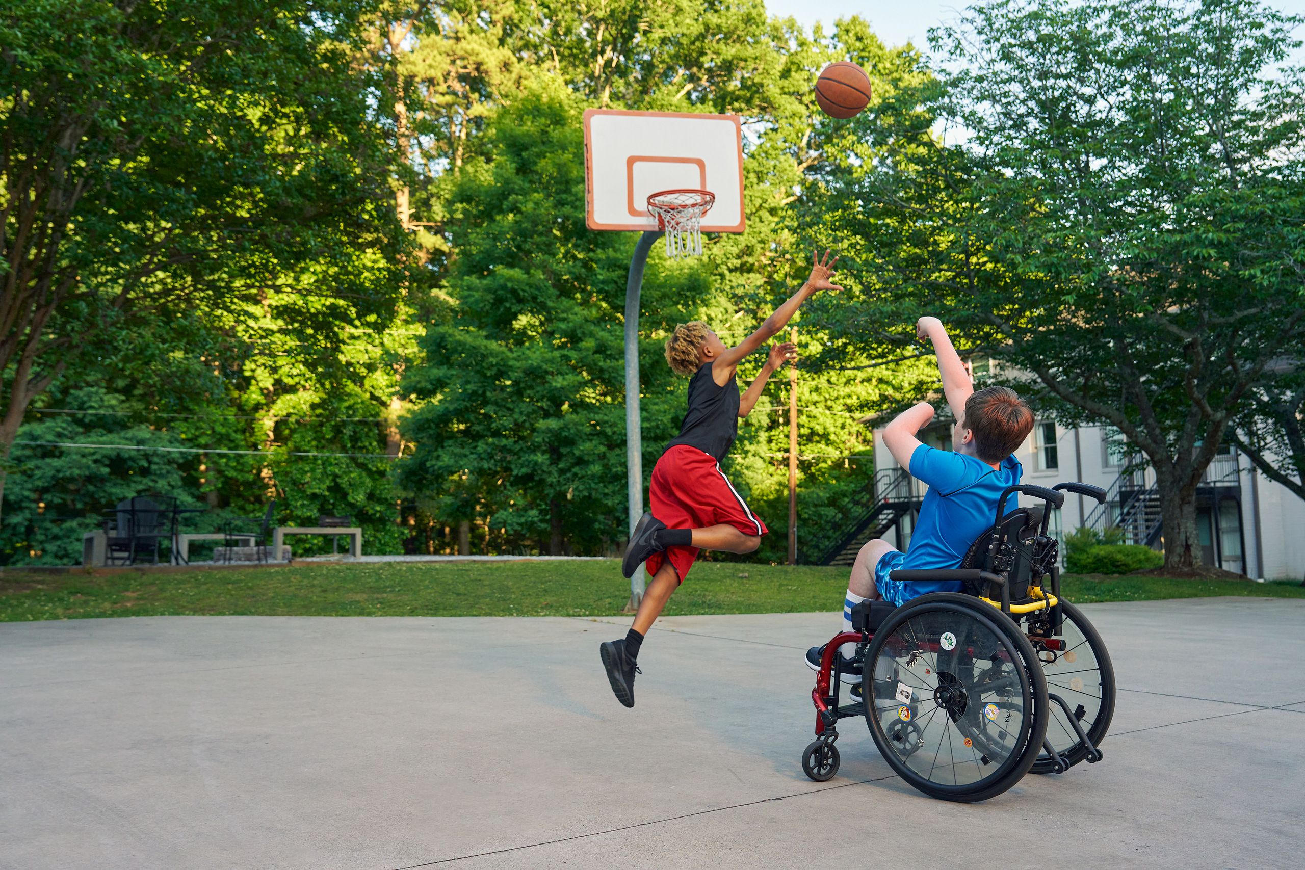 Two teens play basketball at a neighborhood park. One teen, a white boy seated in a manual wheelchair, shoots the ball. The other teen, a light-skinned African American male, jumps forward with arms outstretched, defending the shot. The teen seated in a manual wheelchair has short brown hair and is wearing blue athletic wear. The other teen is wearing black and red athletic wear.