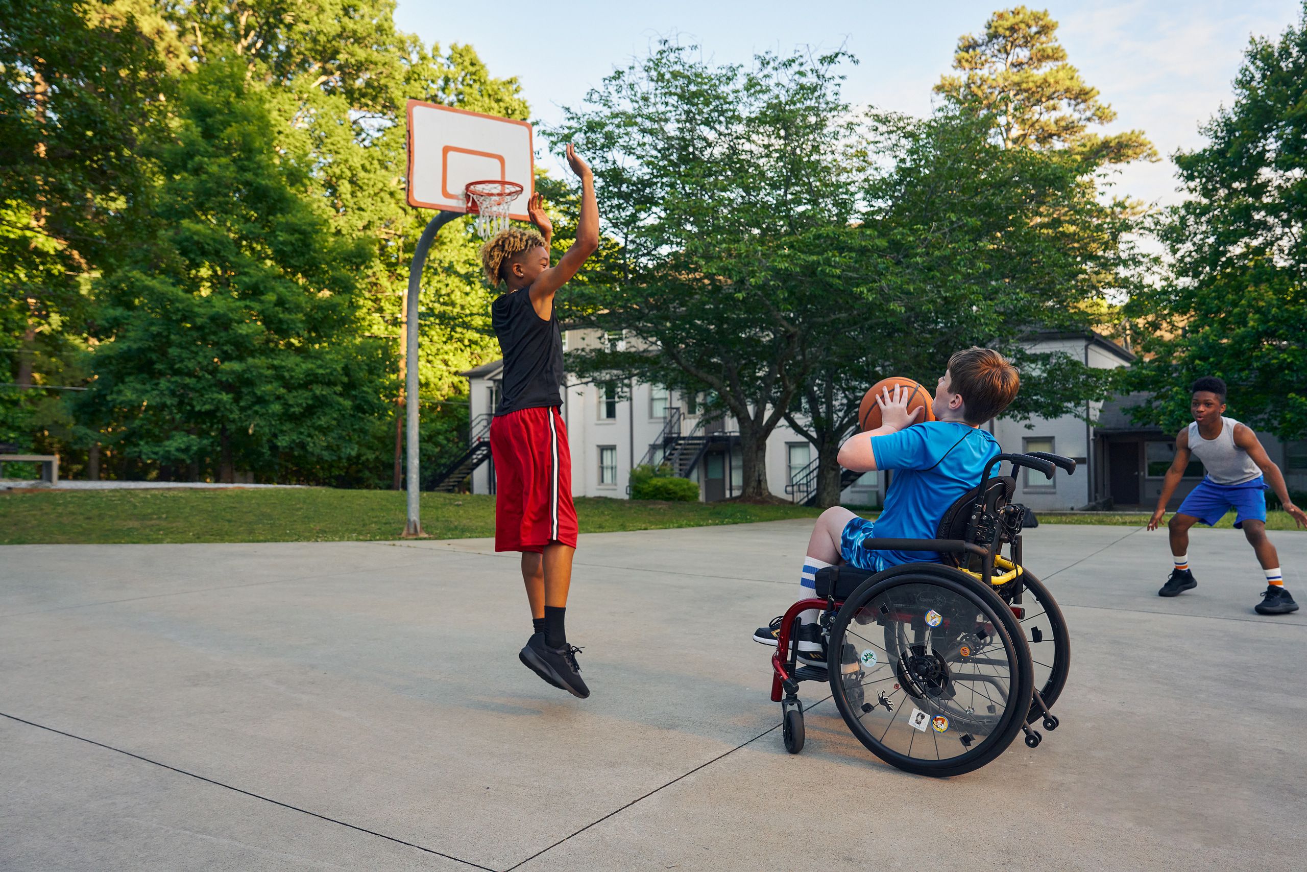 Three teens play basketball at a neighborhood park. One teen, a white boy seated in a manual wheelchair, holds the ball, ready to shoot. A second teen, a light-skinned African American male, jumps forward with arms outstretched, ready to defend the shot. A third teen, a Black boy, crouches, ready to receive a pass. The teen seated in a manual wheelchair has short brown hair and is wearing blue athletic wear. The second teen defending has curly brown to blonde hair is wearing black and red athletic wear. The third teen has short natural black hair styled high on top and is wearing gray and blue athletic wear.