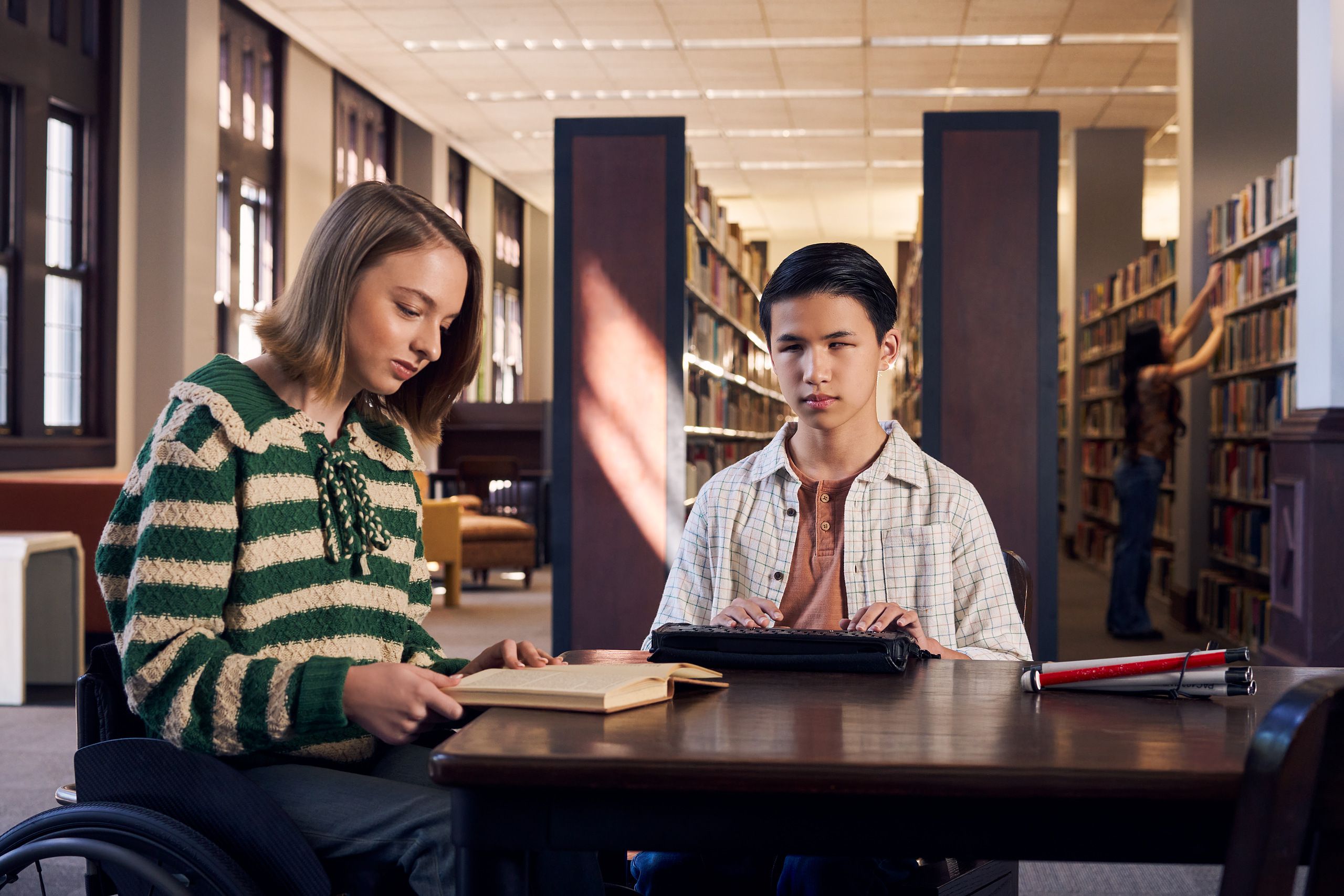 A white teenage girl seated in a manual wheelchair reads a book with focus at a table in a library. Next to her, a Chinese teen, who is a blind cane user, reads on a refreshable braille display. His white cane is collapsed and is placed the table next to him. The teen girl seated in a manual wheelchair has a dark blonde bob and is wearing a green and white striped knit shirt and jeans. The teen boy has slicked back black hair and a friendly smile with braces. He is wearing a rust colored shirt under a white plaid button down shirt.