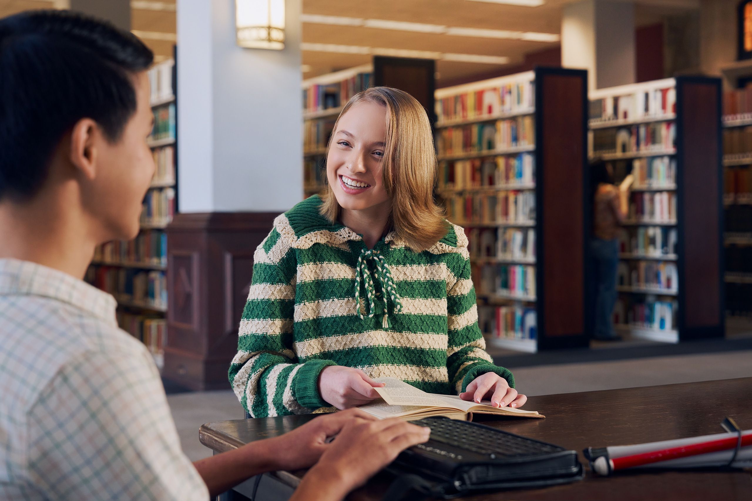 A white teenage girl seated in a manual wheelchair smiles brightly at a friend and holds a book open at a table in a library. Her friend, a Chinese teen, who is a blind cane user, has a refreshable braille display in front of him. His white cane is collapsed and is placed the table next to him. The teen girl has a dark blonde bob and is wearing a green and white striped knit shirt and jeans. The teen boy has slicked back black hair and is wearing a white plaid button down shirt.