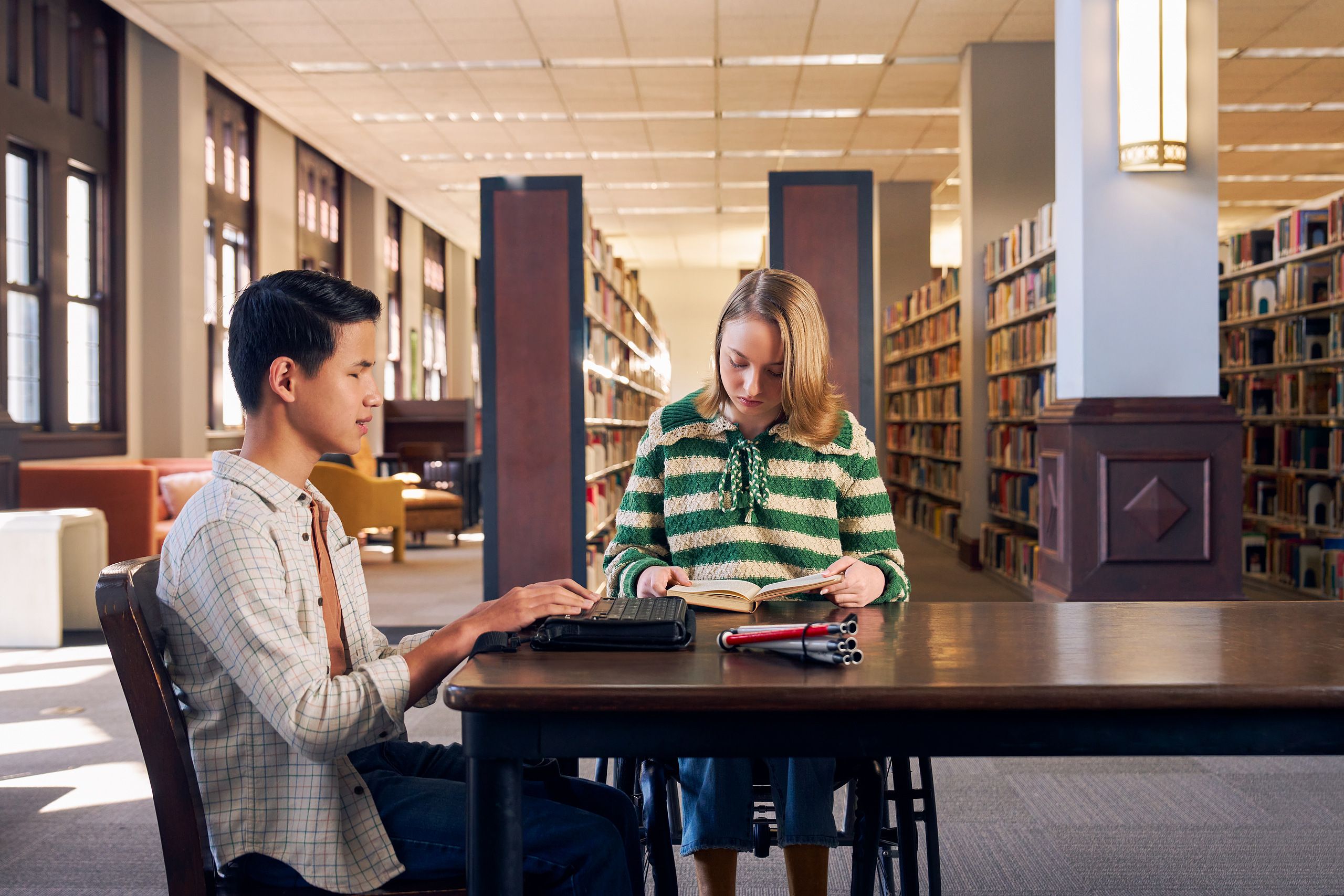 Two teenage friends read at a table in a library. One teen, a Chinese teen, who is a blind cane user, reads on a refreshable braille display. His white cane is collapsed and is placed the table next to him. The other teen, a white teenage girl seated in a manual wheelchair, is seated in a manual wheelchair and holds a hardcover book open. The teen girl has a dark blonde bob and is wearing a green and white striped knit shirt and jeans. The teen boy has slicked back black hair and is wearing a white plaid button down shirt.