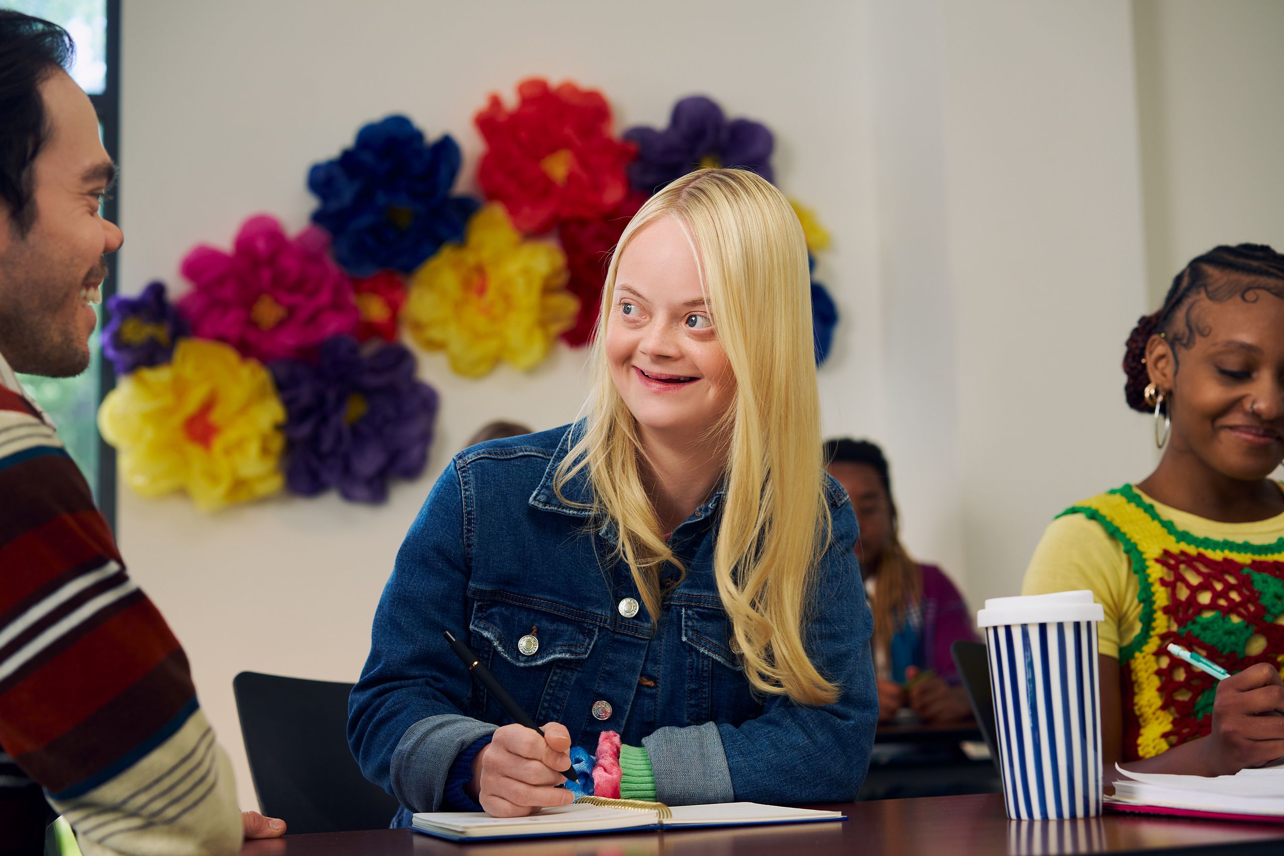 A caucasian college student with Down Syndrome sits at a lecture table, smiles, and chats with her professor, who is standing next to her. She has long blonde hair, blue eyes, and is wearing a jean hacket and several bright scrunchies on her wrist. Another student next to her, a Black woman with braided hair pulled into a bun, writes in a notebook. A large flower sculpture hangs on the wall behind her. Her professor, a white man with medium brown hair and light brown eyes who has dwarfism, smiles and leans on the students' lecture table.