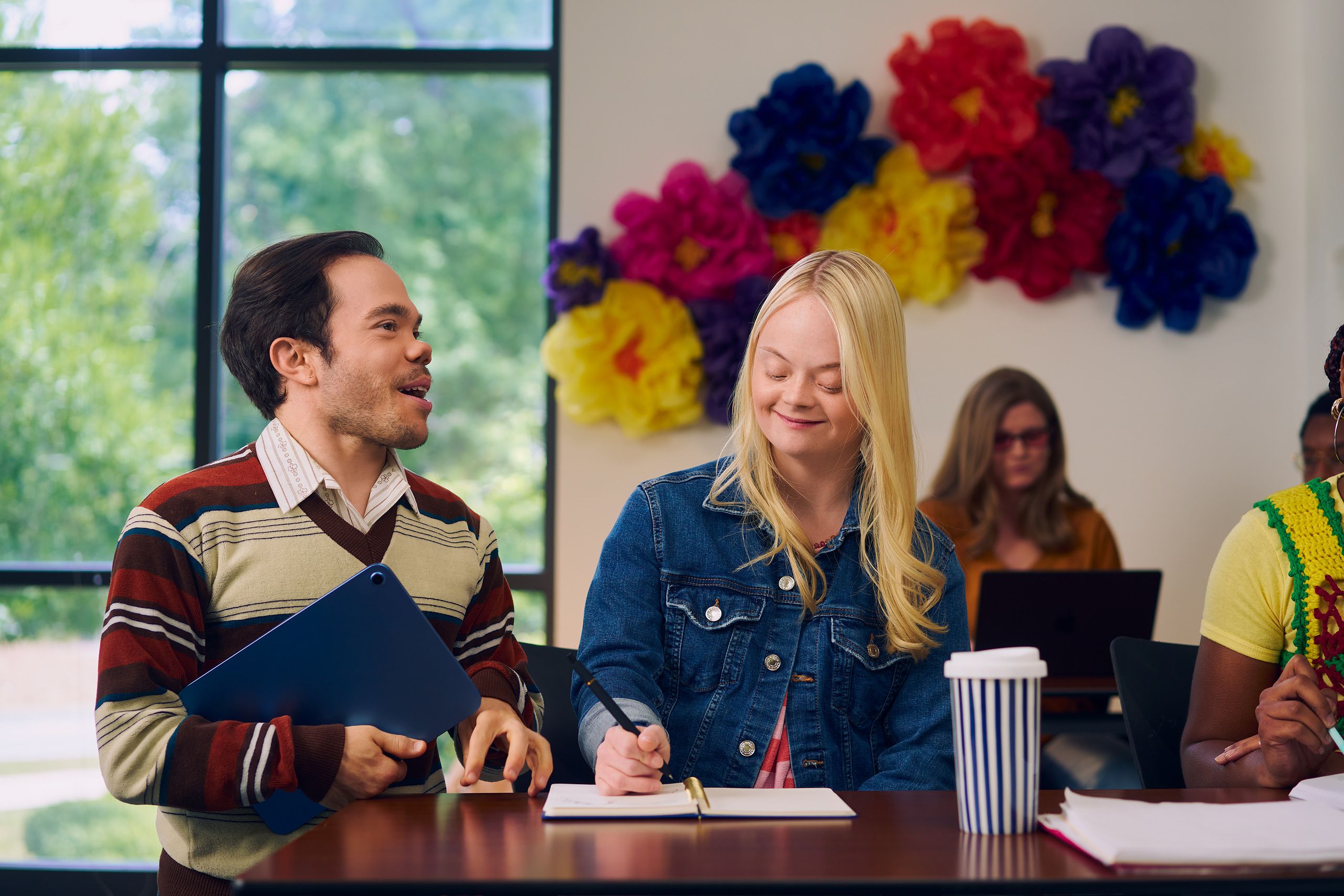 A caucasian college student with Down Syndrome sits at a lecture table and takes notes while her professor, who is standing next to her, chats with her. She has long blonde hair, blue eyes, and is wearing a jean hacket and several bright scrunchies on her wrist. Another student behind her, a  white woman wearing glasses with red lenses, types on a laptop. A large flower sculpture hangs on the wall behind the desks. Her professor, a white man with medium brown hair and light brown eyes who has dwarfism, smiles and holds a tablet in his hand. He has short brown hair and is wearing a red striped sweater, a collared shirt, and dark dress pants.