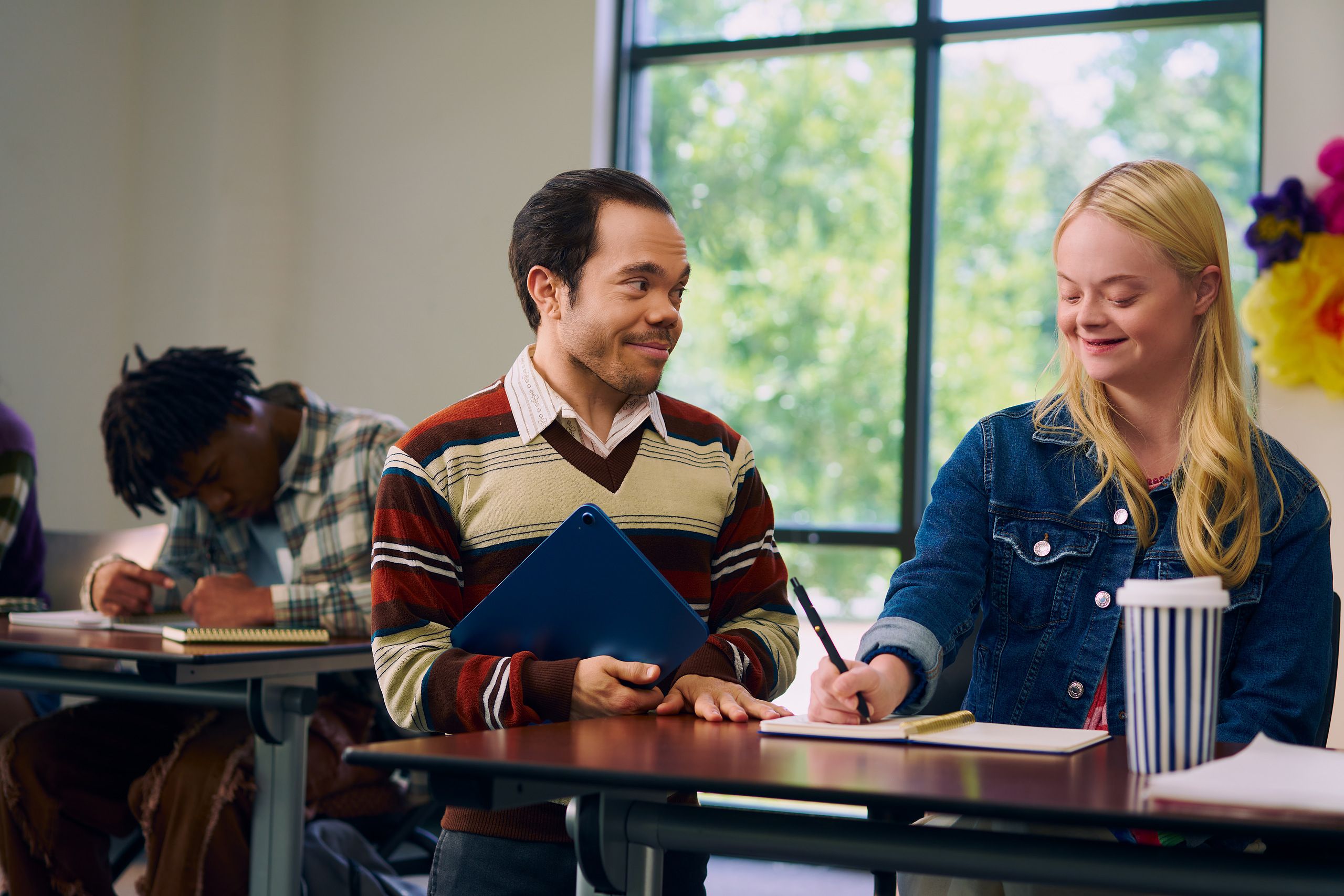 A caucasian college student with Down Syndrome sits at a lecture table and takes notes while her professor, who is standing next to her, chats with her. She has long blonde hair, blue eyes, and is wearing a jean hacket and several bright scrunchies on her wrist. Her professor, a white man with medium brown hair and light brown eyes who has dwarfism, smiles and holds a tablet in his hand. He has short locs and is wearing a red striped sweater, a collared shirt, and dark dress pants. At another table, a Black man with short hair takes notes with focus. A large window with trees showing is at the back of the classroom.