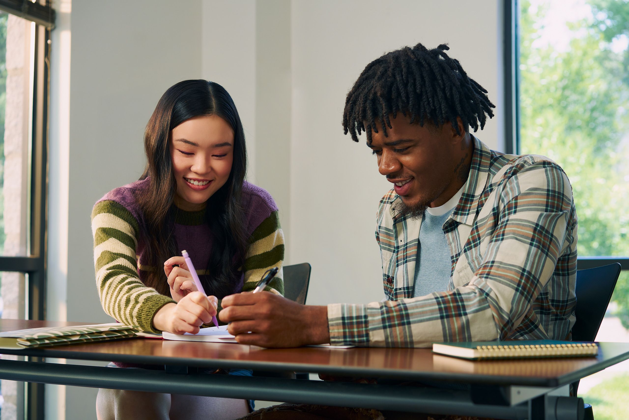Two college students smile and write in a worksheet together at a lecture table. One student, a Black man, has short locs and is wearing a white plaid shirt and a blue knit shirt underneath. The other student, an East Asian young woman, has long curled hair and is wearing a purple, green, and tan striped sweater.