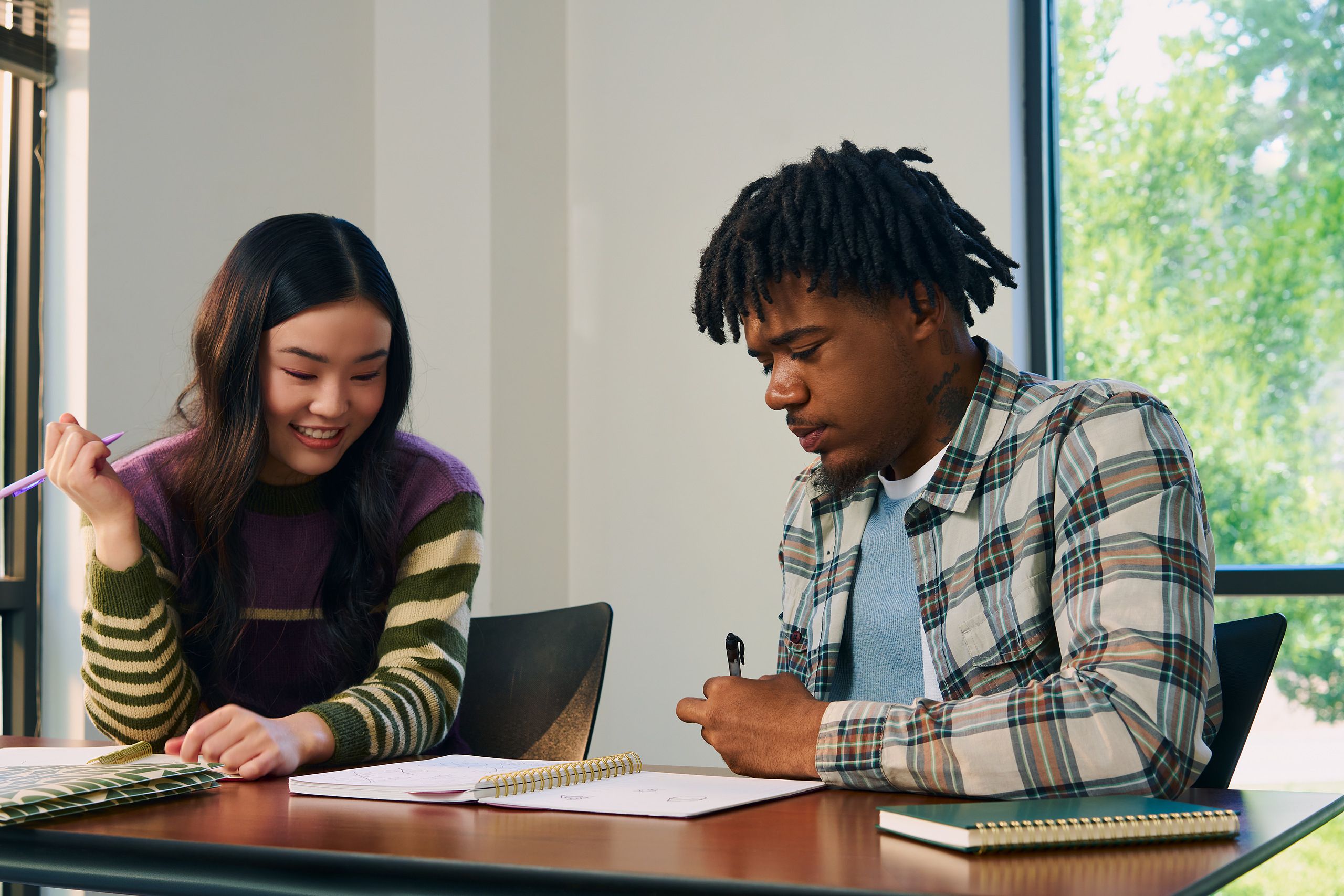 Two college students study notes in an open notebook together at a lecture table. They are both holding pencils. One student, a Black man with short locs is wearing a white plaid shirt and a blue knit shirt underneath. The other student, an East Asian young woman, has long curled hair and is wearing a purple, green, and tan striped sweater.