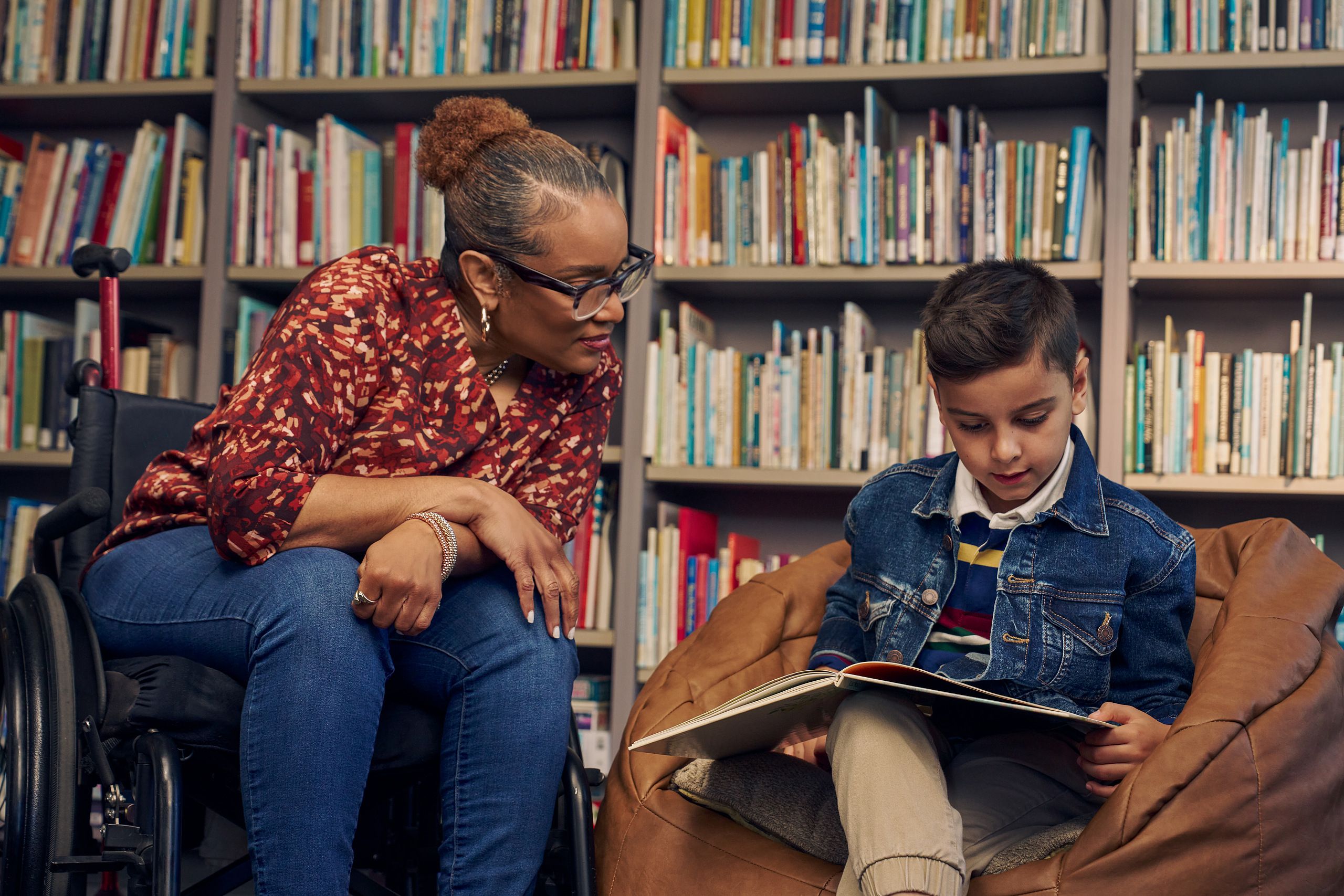 An African American teacher seated in a manual wheelchair watches a young student sitting in a bean bag chair read a picture book. The teacher has medium brown skin, reddish brown hair pulled into a short natural ponytail, and is wearing reading glasses, a red patterned blouse, and jeans. The young student is a Hispanic and Mediterranean-American boy with short brown hair. He has olive skin and is wearing a blue striped shirt, a jean jacket, and khaki pants. A wall of children's books are behind them.
