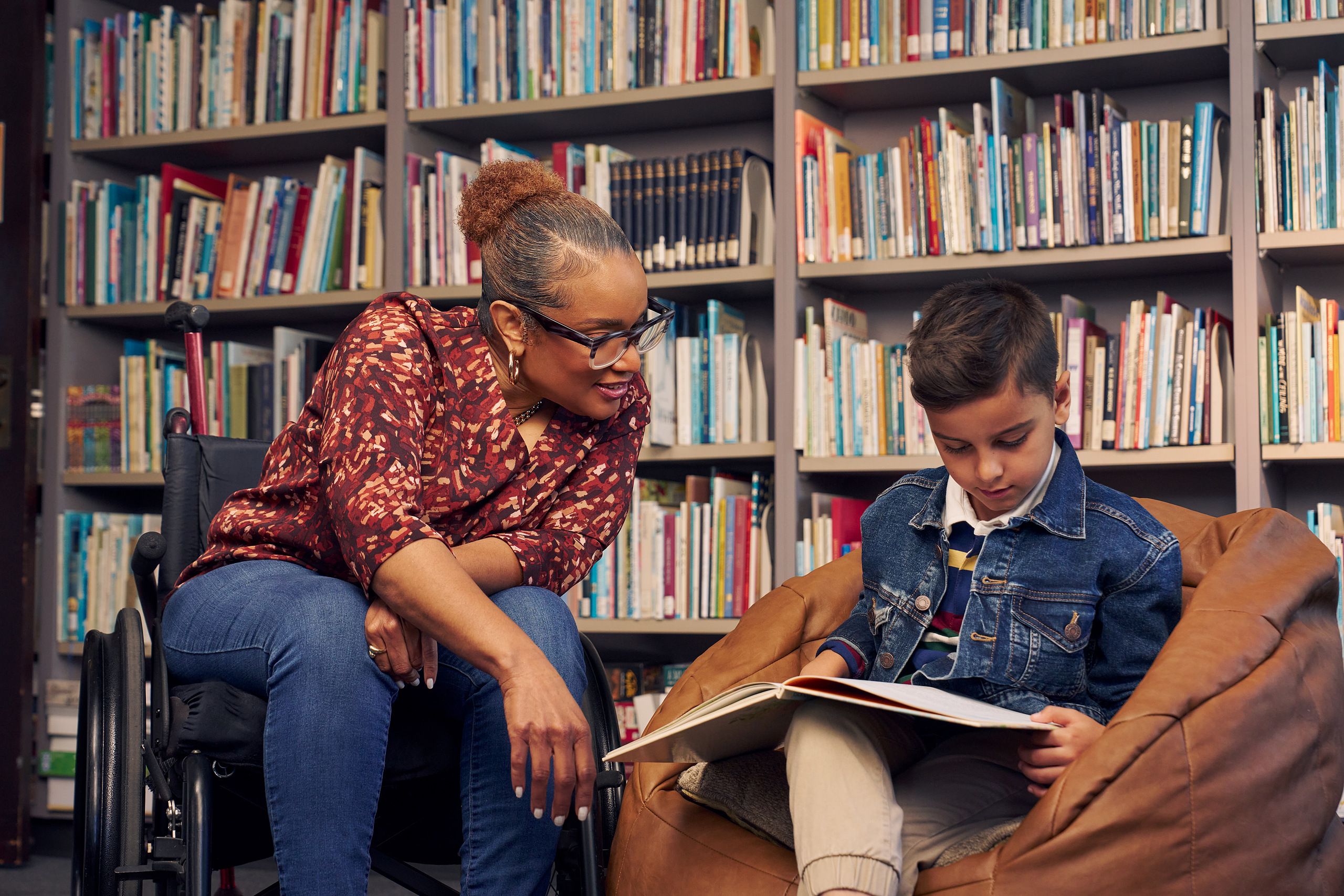 An African American teacher seated in a manual wheelchair smiles and watches a young student sitting in a bean bag chair read a picture book. The teacher has medium brown skin, reddish brown hair pulled into a short natural ponytail, and is wearing reading glasses, a red patterned blouse, and jeans. The young student is a Hispanic and Mediterranean-American boy with short brown hair. He has olive skin and is wearing a blue striped shirt, a jean jacket, and khaki pants. A wall of children's books are behind them.