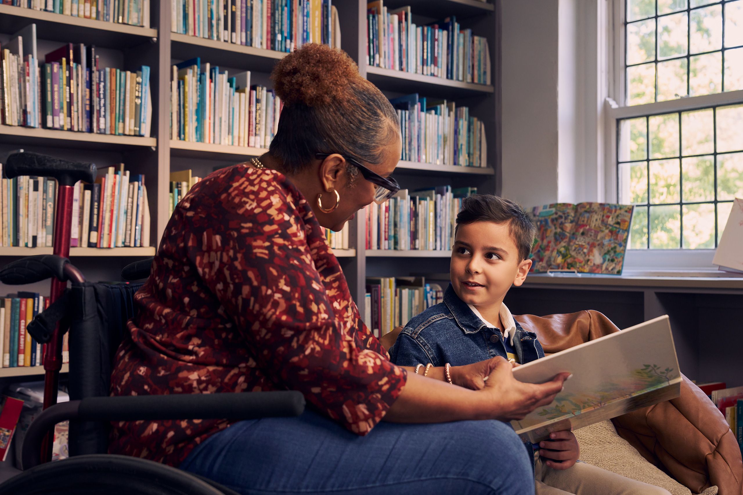 A young boy sitting in a bean bag chair looks up at his teacher, an African American woman seated in a manual wheelchair. They are both holding a picture book. The teacher has a red walking cane fastened to the back of her wheelchair. She has medium brown skin, reddish brown hair pulled into a short natural ponytail, and is wearing reading glasses, a red patterned blouse, and jeans. The young student is a Hispanic and Mediterranean-American boy with short brown hair. He has olive skin and is wearing a blue striped shirt, a jean jacket, and khaki pants. A wall of children's books are behind them.