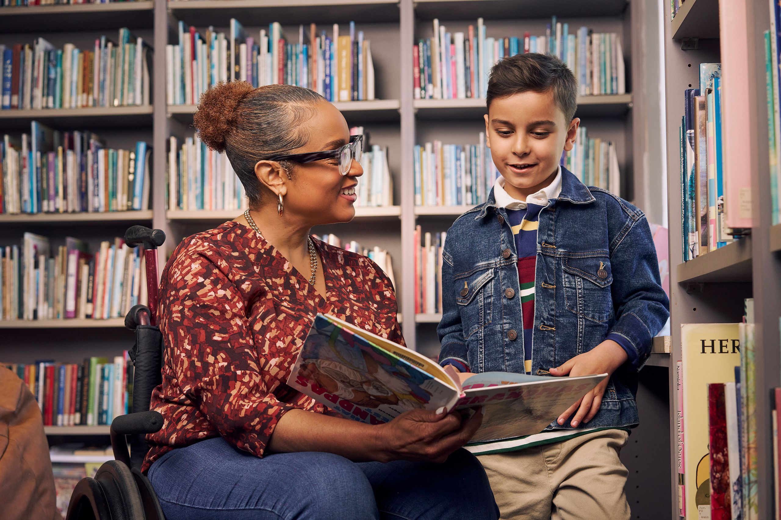 An African American teacher seated in a manual wheelchair smiles and holds a picture book open while her young student reads out loud. The teacher has medium brown skin, reddish brown hair pulled into a short natural ponytail, and is wearing reading glasses, a red patterned blouse, and jeans. The young student is a Hispanic and Mediterranean-American boy with short brown hair. He has olive skin and is wearing a blue striped shirt, a jean jacket, and khaki pants. A wall of children's books are behind them.