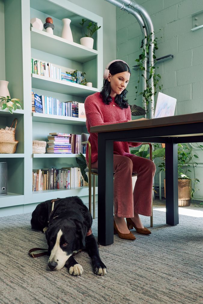 A young pale white blind woman is sitting at a desk typing on a laptop computer in a modern coworking office space. A black and white guide dog wearing a guide harness is lying on the floor near her feet. The woman has shiny black hair styled in waves and is wearing over the ear headphones, a chunky knitted pink sweater, dusky pink flared pants, and taupe pointed toe suede heels. The coworking office space is painted a warm mint green and features a large bookcase with several books, plants, a rattan basket, and clay vases.