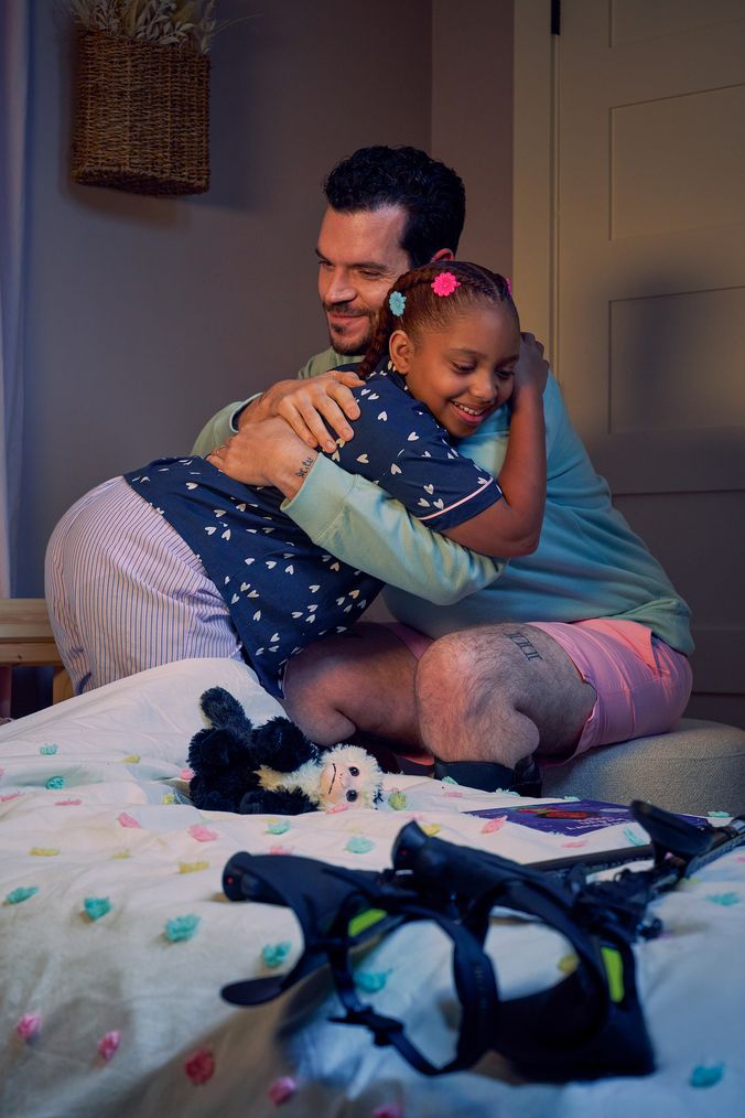 A father and daughter smile and embrace in her bedroom at night. The father is a white man with Cerebral Palsy who wears leg braces. His black forearm crutches lie on the foot of the daughter's bed. The daughter is a Black multiracial American girl with thick braided red hair adorned with two flower barrettes and is wearing blue patterned pajamas. A Capuchin monkey plushie lays on her bed.