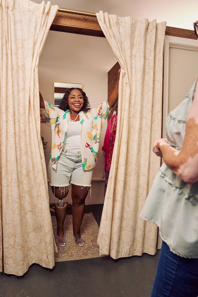 A Black woman with a prosthetic leg opens up the curtains of a dressing room of a boutique with a big smile on her face. She is wearing a patterned white blouse, a white tank, jean cut off shorts, and jelly sandals. She looks at the shopkeeper, partially off-camera. Several high end garments are on hooks in the dressing room. The shopkeeper is a Black woman with Vitiligo. She has a short, curly bob and is wearing glasses, a flowy blouse, and jeans.