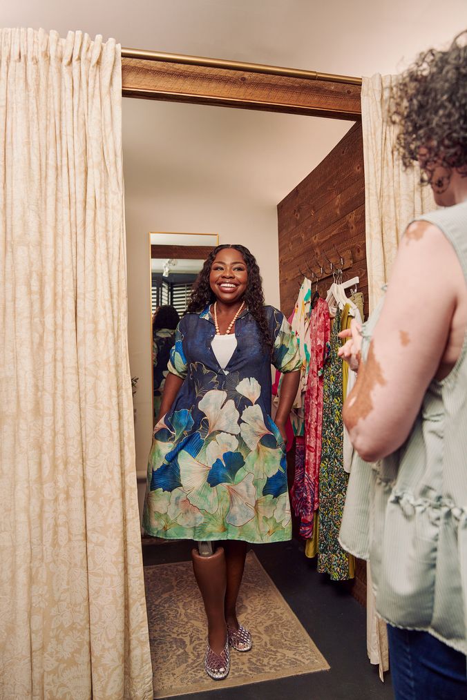 A Black woman with a prosthetic leg poses in a dressing room of a boutique with a big smile on her face. She is wearing a blue patterned collared dress and jelly sandals. She has her hands in the pockets of the dress and looks at the shopkeeper, partially off-camera. Several high end garments are on hooks in the dressing room. The shopkeeper is a Black woman with Vitiligo. She has a short, curly bob and is wearing glasses, a flowy blouse, and jeans.