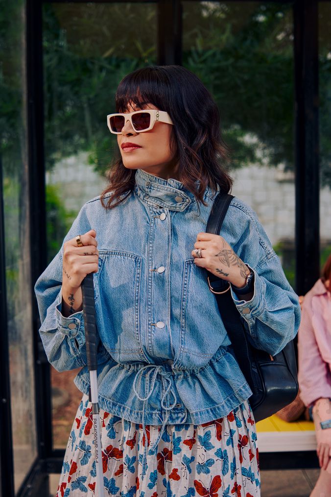 An Asian Filipino woman with Retinitis Pigmentosa and Usher Syndrome stands at a covered bus stop. She is wearing white sunglasses and is holding her white cane with her tattooed hands. She has on a jean jacket, a butterfly patterned skirt, and a black backpack.