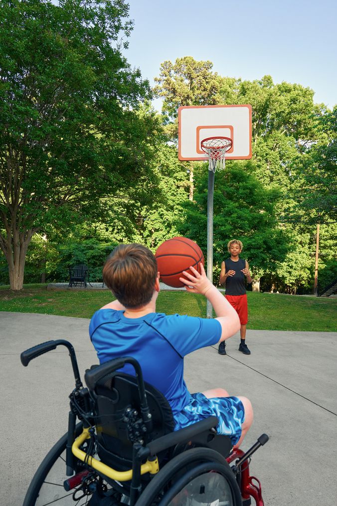 Two teens play basketball at an outdoor park. One of the friends, a white boy seated in a manual wheelchair, passes the ball to the other, a light-skinned African American male, who is standing near the hoop. The teen seated in the manual wheelchair has short brown hair is wearing blue athletic wear and faces away from the camera. The African American teen has curly brown to blonde hair and is wearing black and blue athletic wear.