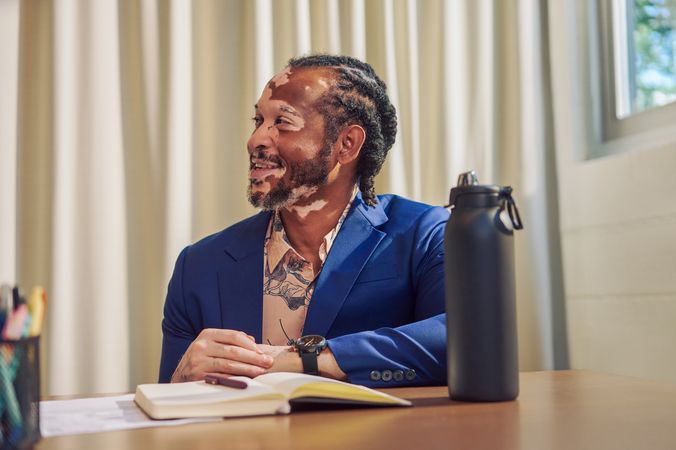 An African American man with olive and porcelain skin and Non-Segmental Vitiligo is seated at a conference room table, smiling at a colleague off-camera. A black water bottle is in front of him. He has brown barrel-twist dreadlocks, wears a navy blue suit with light pink and floral blue-patterned dress shirt. His brown eyes and black watch offset his black goatee.