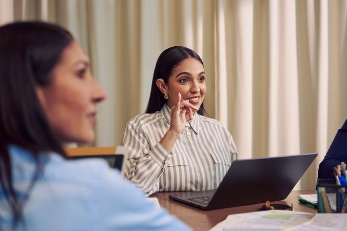 A Deaf Latina Puerto Rican woman uses American Sign Language while seated at a conference room table with two colleagues. She has long deep brown hair and is wearing a striped business shirt and hoop earrings. A laptop and business planning documents are in front of her.